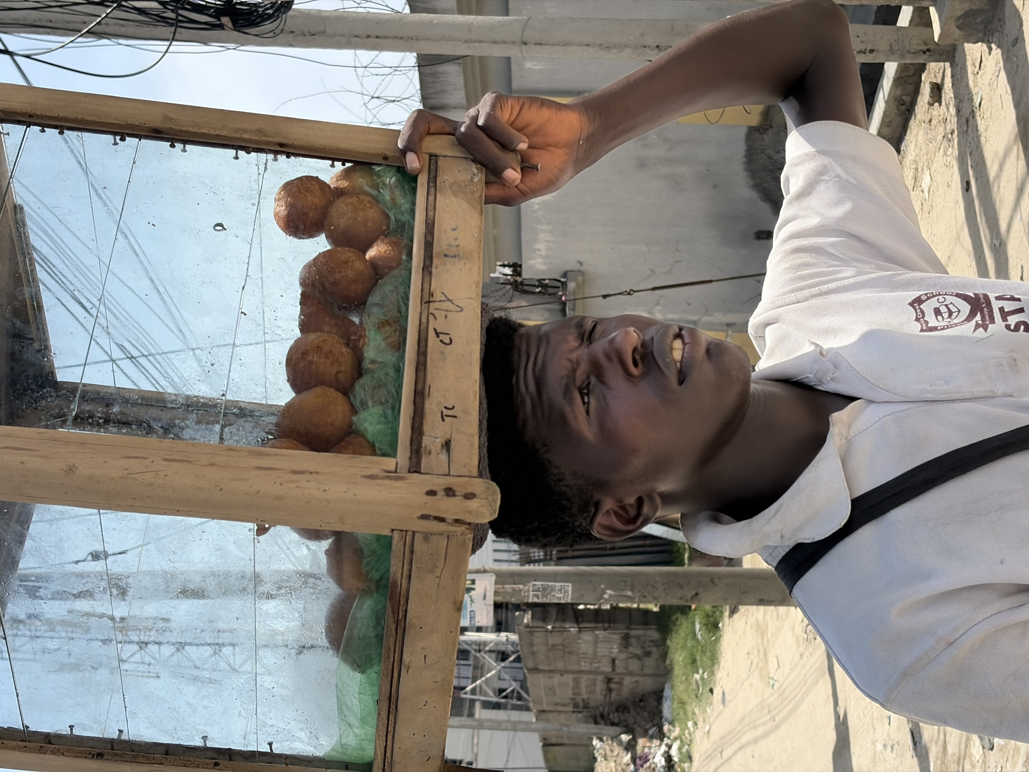 A medium shot depicts an Igbo man standing in what appears to be Igboefon, Nigeria, based on the writing on the food cart. He's next to a food cart constructed of wood and glass, containing a variety of round, brown, fried dough balls, possibly Akara or Puff-Puff, lying on a green plastic surface. The man wears a white button-down shirt with a logo in the upper right corner and a black strap over his left shoulder. He has short, curly hair and slightly prominent teeth. The background features buildings, an electric pole, and trash on the ground. The camera angle is slightly tilted.