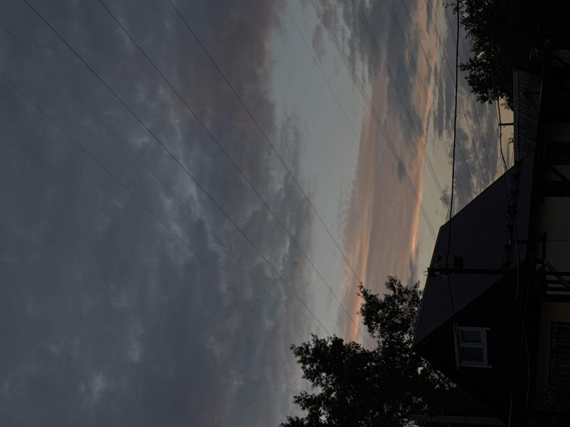 The image shows a dark silhouette of a house and trees against a twilight sky. The sky is filled with clouds, with streaks of pink and orange visible, suggesting the sun is setting or has recently set. Several power lines crisscross the sky. The house appears to be a two-story structure with a dark, sloped roof and a visible window. The overall scene is dim and atmospheric, indicative of dusk. There are no people visible in the image. The location is the city of Chaikovsky, Russia.