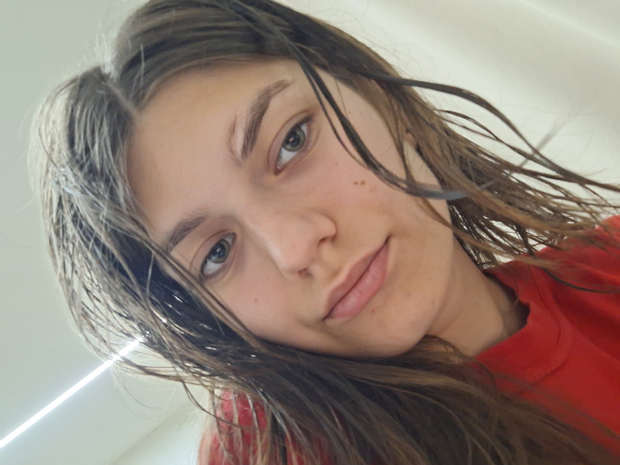 A close-up, slightly angled, selfie-style shot shows a young woman with damp, long, brown hair. Her skin appears fair, with a few freckles visible on her face. She has light eyes, seemingly green or hazel. The subject is wearing a red shirt. The background is out of focus, showing a plain, light-colored wall with a fluorescent light fixture. There is no clear indication of the location being Panagyurishte, Bulgaria, or any specific city context in the image.