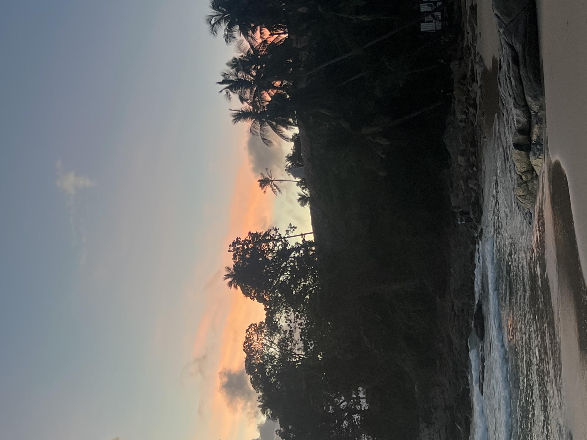 A beach scene in Choeng Thale, Thailand, at dusk. The sky transitions from a pale blue at the top to hues of pink and orange near the horizon, with scattered clouds. Silhouetted palm trees and dense foliage line a rocky incline overlooking the beach. The ocean has gentle waves breaking on the shore, which is composed of sand and large rocks. In the distance, a few small structures are visible among the trees on the right side of the frame. The lighting suggests the sun has recently set.