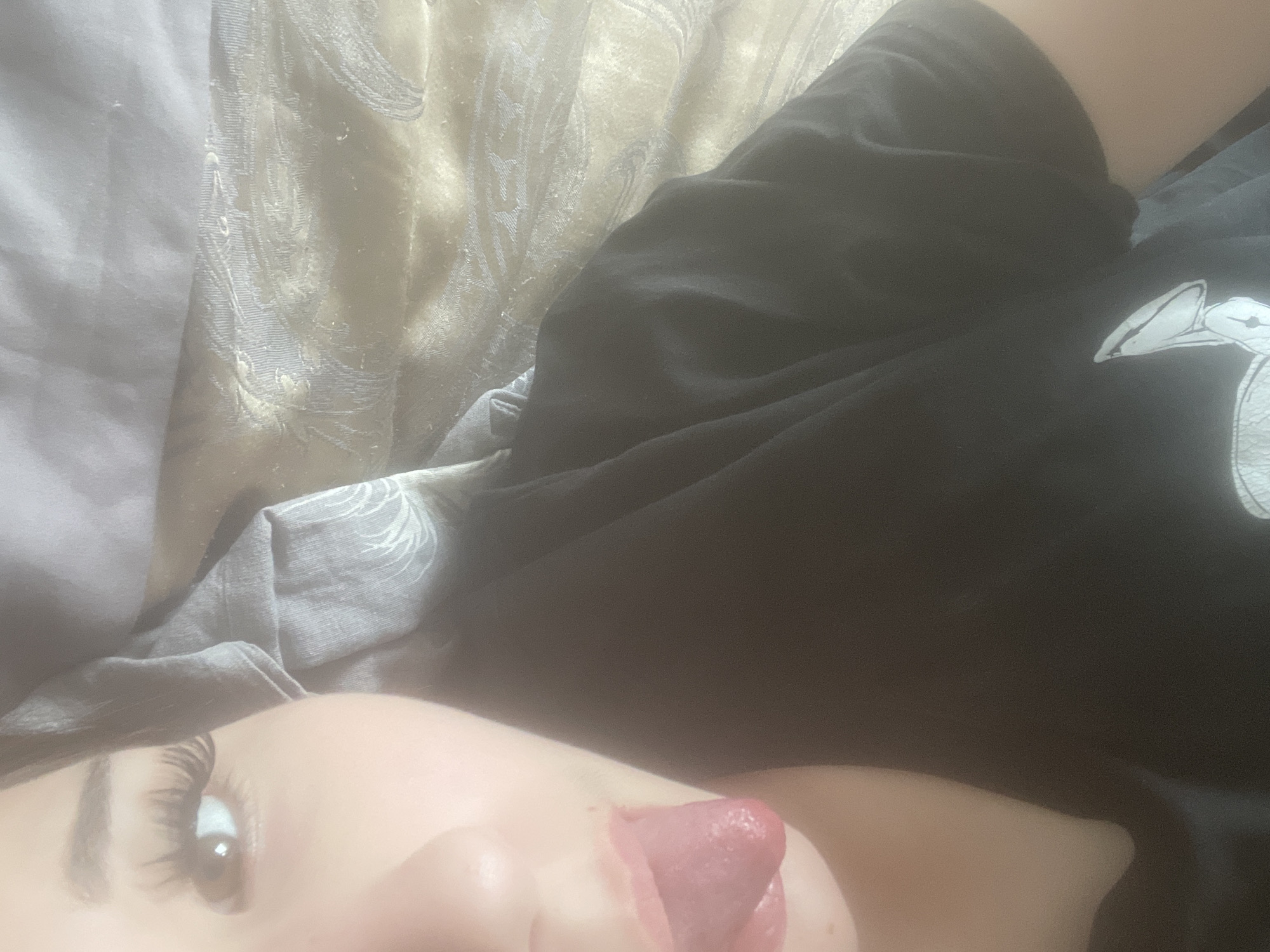 An indoor, close-up image captures a portion of a person's face and upper body, lying down in what appears to be a bedroom setting. The individual, with fair skin, is visible from their right eye and eyebrow to their mouth, where their tongue is playfully extended. Their eye features prominent, dark eyelashes and a defined eyebrow.
They are wearing a dark-colored garment, likely a t-shirt or top, which partially displays a white graphic design on the left side, resembling a stylized animal head. A segment of the person's bare shoulder or upper arm is also visible. Behind and beside them are various fabrics: a light-colored, textured material with intricate, swirling patterns, possibly a pillow or blanket, and a plain lighter-toned fabric. Another folded grey fabric is also present near their head.
The lighting in the image is soft and somewhat diffused, creating a slightly hazy effect, and contributing to a relaxed atmosphere. There is no visible text in the image. The scene offers no specific visual cues to indicate the location as Sinyavino, Russia.