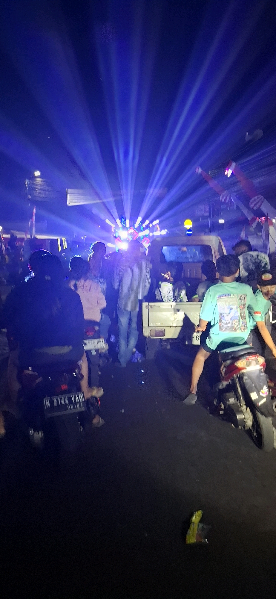 The image captures a nighttime scene in Prigen, Indonesia, characterized by vibrant blue stage lights that illuminate a gathering of people. The lights project outwards in distinct beams, creating a dramatic effect. In the foreground, several individuals are visible, some on motorcycles and others standing. One person in the center wears a striped long-sleeved shirt and blue jeans. To the right, another individual is wearing a light blue t-shirt and dark shorts, seated on a motorcycle. Behind them, a white pickup truck is parked, also illuminated by the stage lights. The ground is asphalt, and there is some scattered debris, including a crumpled yellow and red wrapper. The overall atmosphere appears festive, likely at an outdoor event or performance.