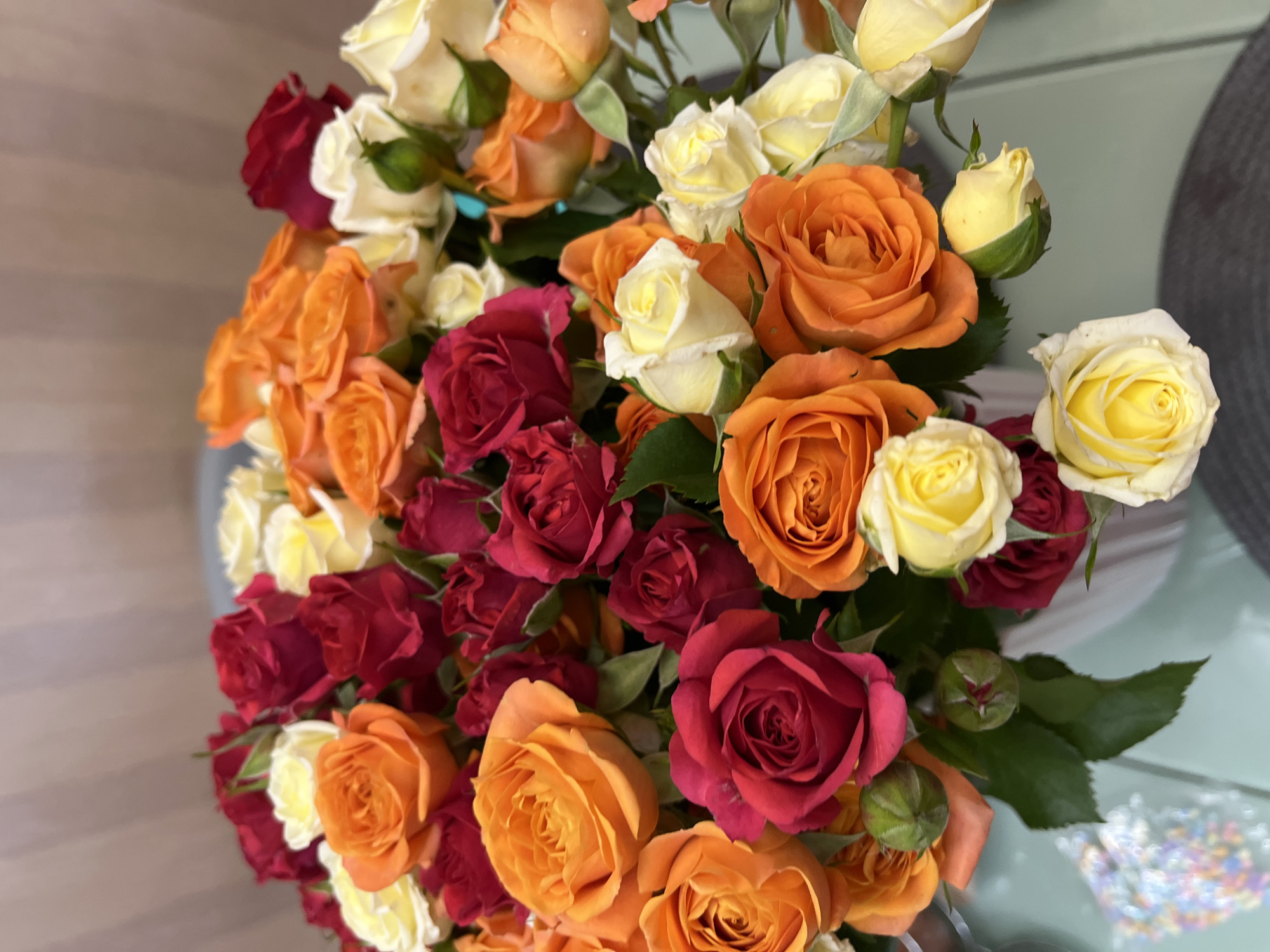 This is a close-up, eye-level shot of a bouquet of roses in a white vase. The roses are a mix of red, orange, and yellow, with some buds visible. The background is a blurred, striped wall. The bouquet appears to be on a light blue surface, possibly a table, with some out-of-focus colorful objects in the lower left corner. The image does not provide any information about the city of Sochi, Russia.
