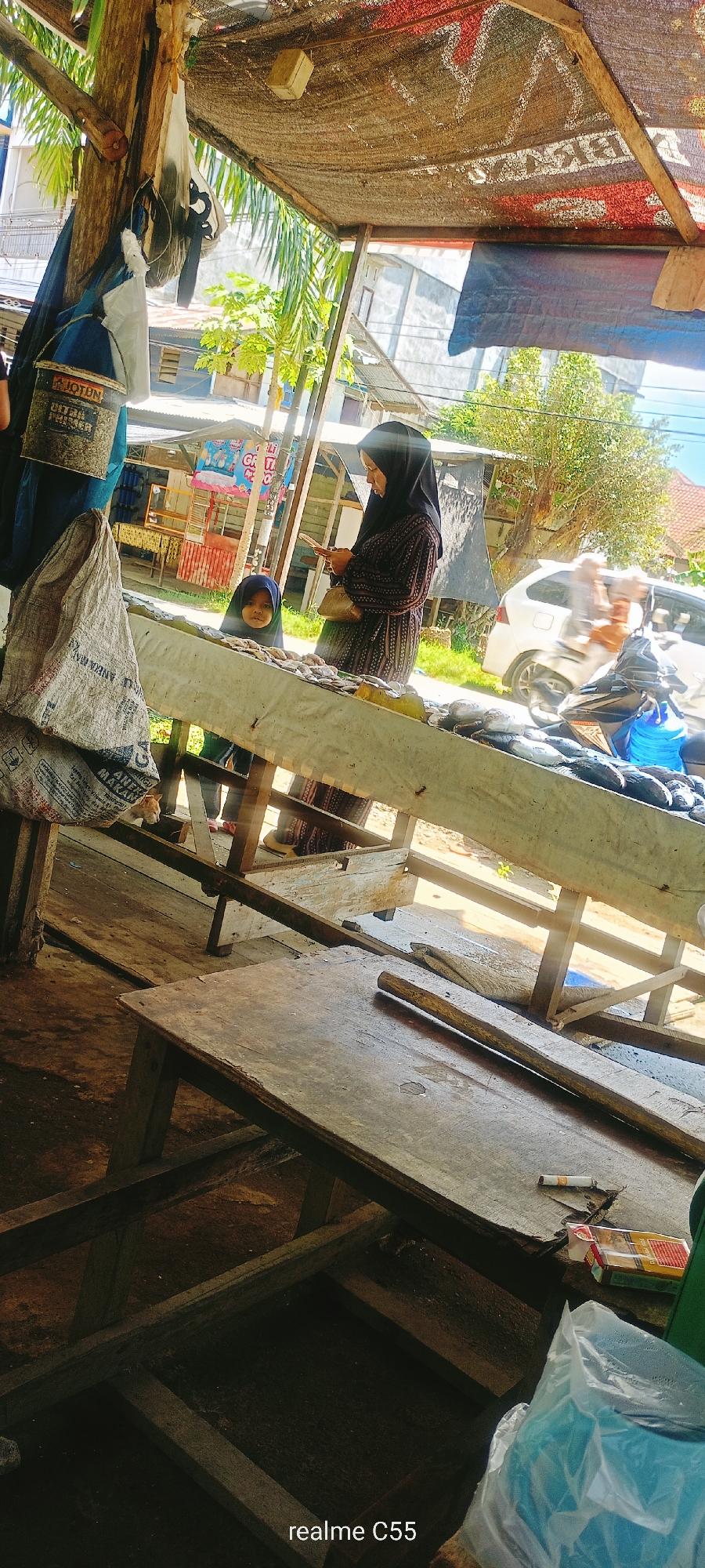 The image depicts a market stall in Lhokseumawe, Indonesia. A woman wearing a black hijab is standing and looking at her phone. To her left, a young child wearing a blue hijab is looking towards the camera. They are standing behind a table displaying a variety of fish for sale. In the foreground, there are two large sacks, and to the left of these sacks, a ginger-colored cat is visible. The stall is made of wood and is covered by a woven material. In the background, there are other market stalls, buildings, and a white car passing by. The overall scene suggests a typical day at a local market.