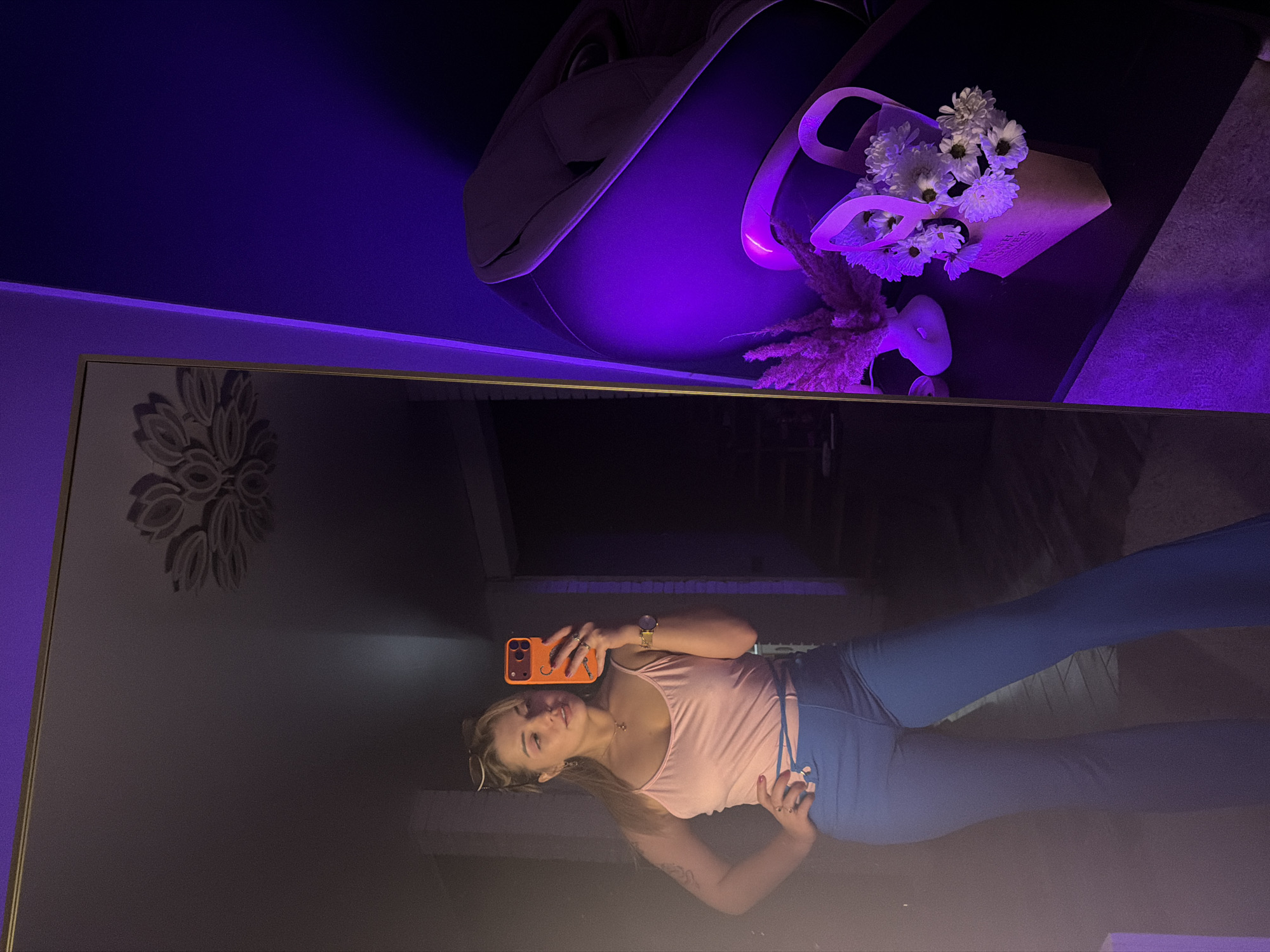 An indoor scene in Большая Шильна, Russia, captured as a mirror selfie. A female individual, appearing to be a young adult with light hair, stands reflected in a large, rectangular mirror with a gold-colored frame. She wears a light pink spaghetti strap tank top, blue high-waisted flared pants with lacing details, sunglasses on her head, a watch on her left wrist, and rings on both hands. A tattoo is visible on her upper left arm. She holds an orange-cased smartphone in her right hand, directed towards the mirror, while her left hand rests on her hip. Her gaze is directed towards the smartphone. Reflected in the upper portion of the mirror is a decorative ceiling fixture. To the right of the mirror, a wall is illuminated with intense purple lighting. On a dark surface against this wall, a small beige paper bag containing white flowers is present, alongside a white, abstractly shaped vase holding reddish-brown dried plant material. A large, dark furniture item, possibly a chair or cabinet, is partially visible in the upper right under the purple light. The background reflected behind the subject is dimly lit, suggesting an interior space.