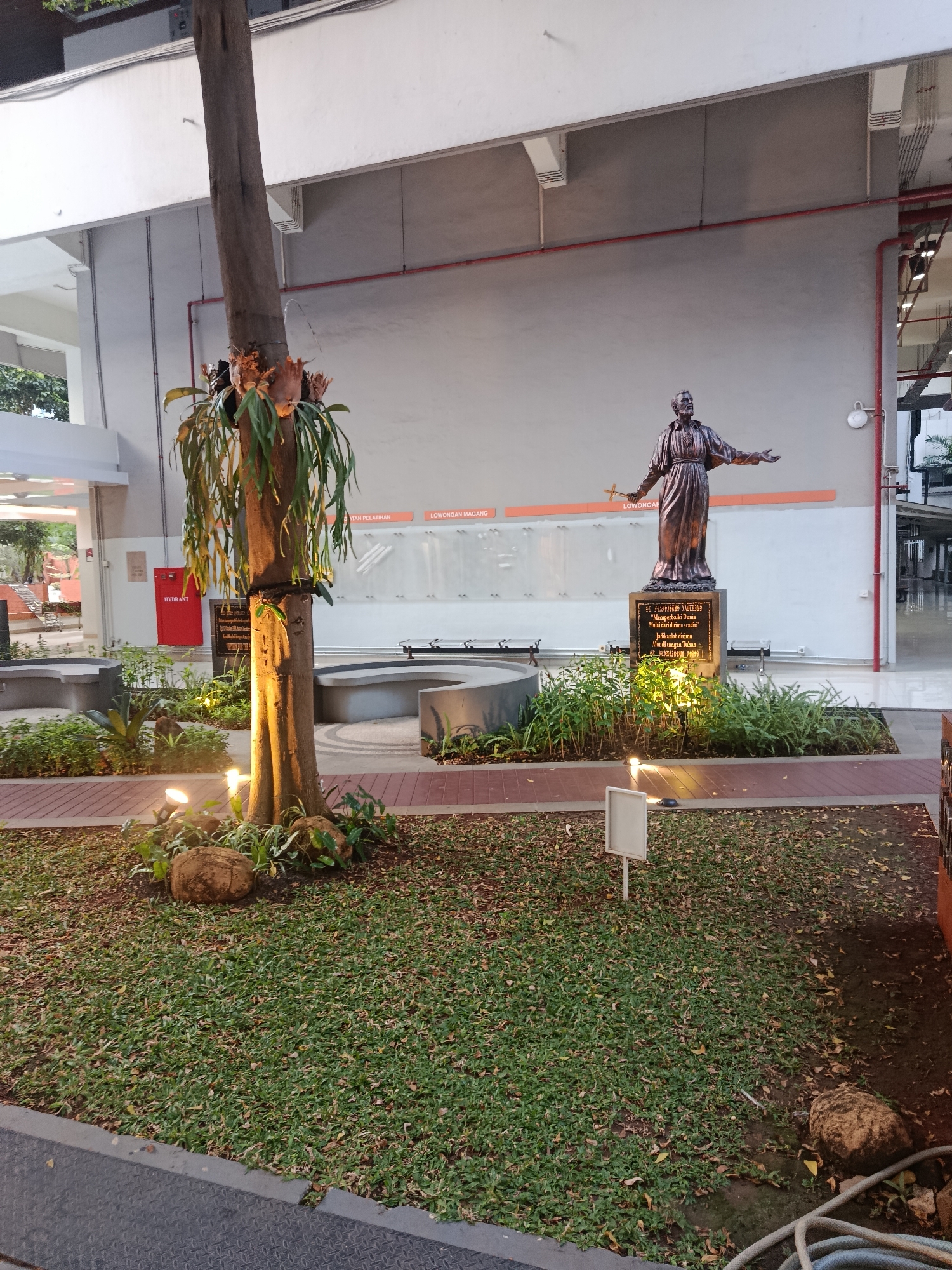 The image depicts an outdoor area, possibly a garden or courtyard, in the city of South Jakarta, Indonesia. A bronze statue of a religious figure, possibly a saint, stands on a pedestal in the center of the scene. The statue is dressed in robes and holds a cross in its outstretched left hand. The pedestal features an inscription, though it is largely illegible in the image.
In front of the statue, there are beds of green foliage and ornamental plants. A large tree with epiphytic plants growing on its trunk is situated to the left of the statue. The tree is illuminated by spotlights, casting warm light on the surrounding area.
The ground is covered with grass and fallen leaves, with some large rocks interspersed. To the left, there is a bright red fire hydrant with the word "HYDRANT" printed on it in white. Behind the statue, a light-colored wall serves as a backdrop, with orange horizontal lines and some indistinct signage.
In the background, architectural elements of a building are visible, including concrete structures, railings, and red pipes. The scene appears to be set during dusk or evening, with artificial lighting illuminating the foreground. There are no people clearly visible in the image.