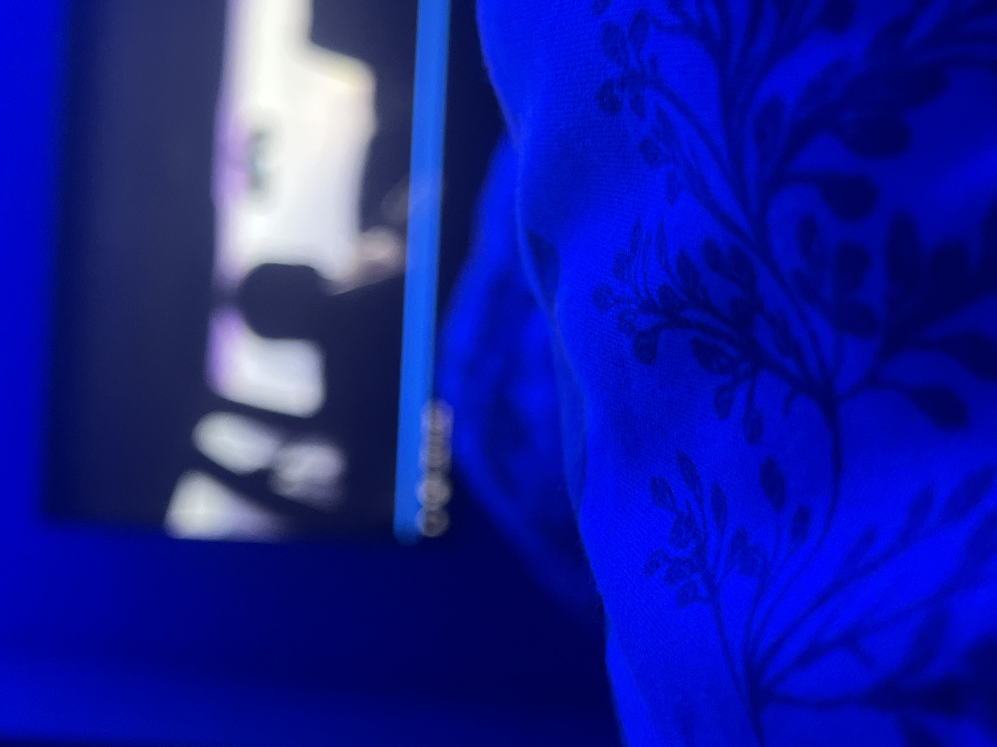 The image depicts a close-up view of a blue fabric with a dark floral pattern, likely a blanket or bedspread. In the background, a blurred screen displays bright, abstract shapes and a dark silhouette of a person's head and shoulders. The lighting is predominantly blue, casting a vibrant glow on the fabric and creating a dim, atmospheric background. The scene appears to be indoors, possibly in a bedroom or a living area. There is no information available to determine the specific location within Vratsa, Bulgaria.