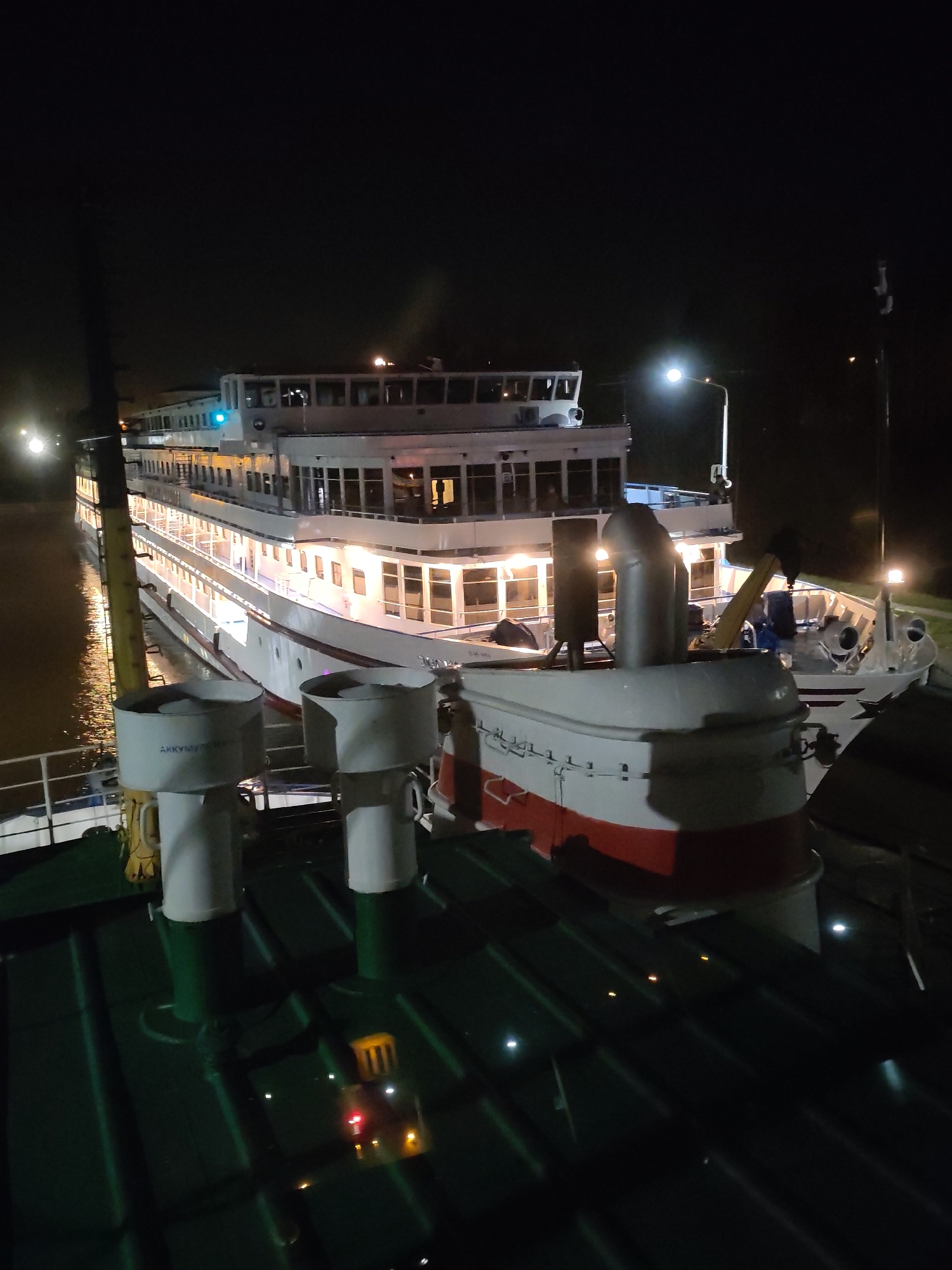 At night, a multi-decked river cruise ship is docked, illuminated with lights. It is situated adjacent to a green-decked vessel. The location is unspecified within the city of Unknown, Unknown. No individuals are visible.