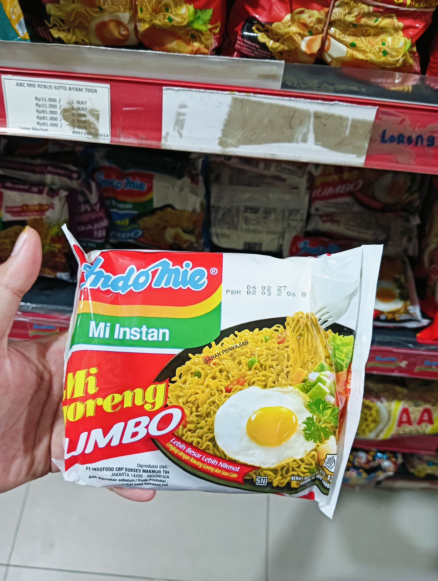 In Batam, Indonesia, a hand is holding a packet of "Indomie Mi Goreng Jumbo" instant noodles. The packet is predominantly red with "Indomie" logo in blue and white, "Mi Instan" in white, and "Mi Goreng JUMBO" in yellow and red. The noodles are depicted on a black plate with a fried egg and garnishes. Behind the packet, other Indomie noodle packets are visible on a store shelf. A price tag for "ABC MIE REBUS SOTO AYAM 70GR" is also visible on the shelf. The environment suggests a retail setting, likely a supermarket or convenience store.