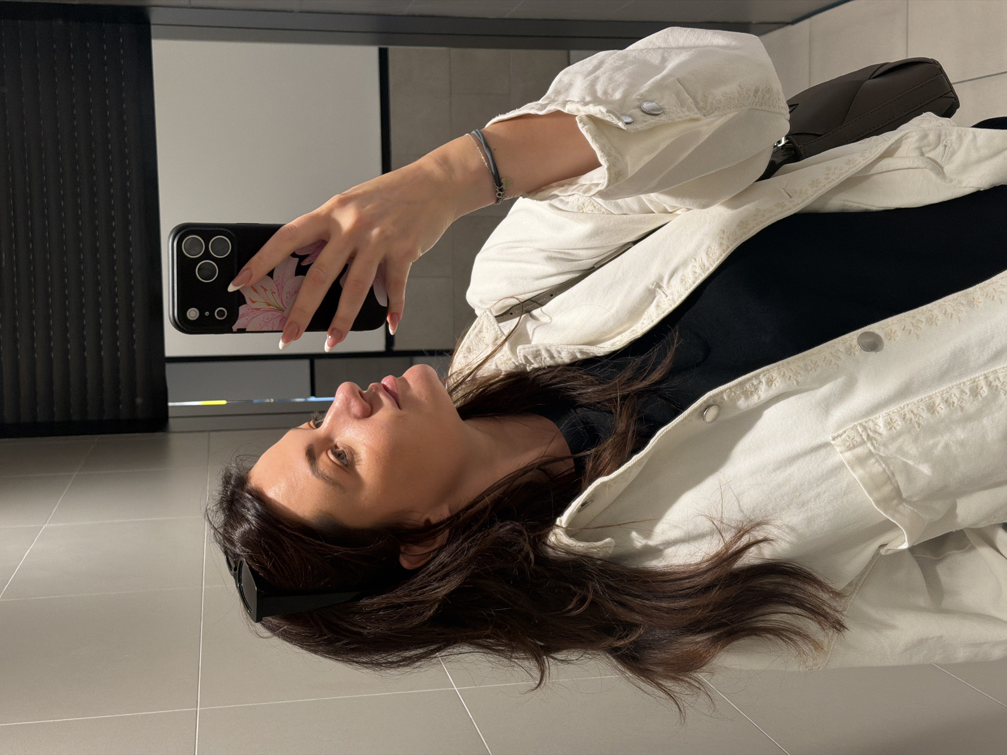A woman is taking a selfie in a room with light gray tile flooring and a white wall. The location may be in Belgrade, Serbia, but there is no definitive evidence in the image. She is holding a smartphone with a floral design on the case, displaying a three-lens camera. The woman has brown hair and fair skin, wearing a black shirt under a white, button-up denim jacket. She has a bracelet on her left wrist. In the background is a dark textured wall and a white surface, which could be a wall or a screen. A dark handbag is partially visible. She is facing the camera at an angle.