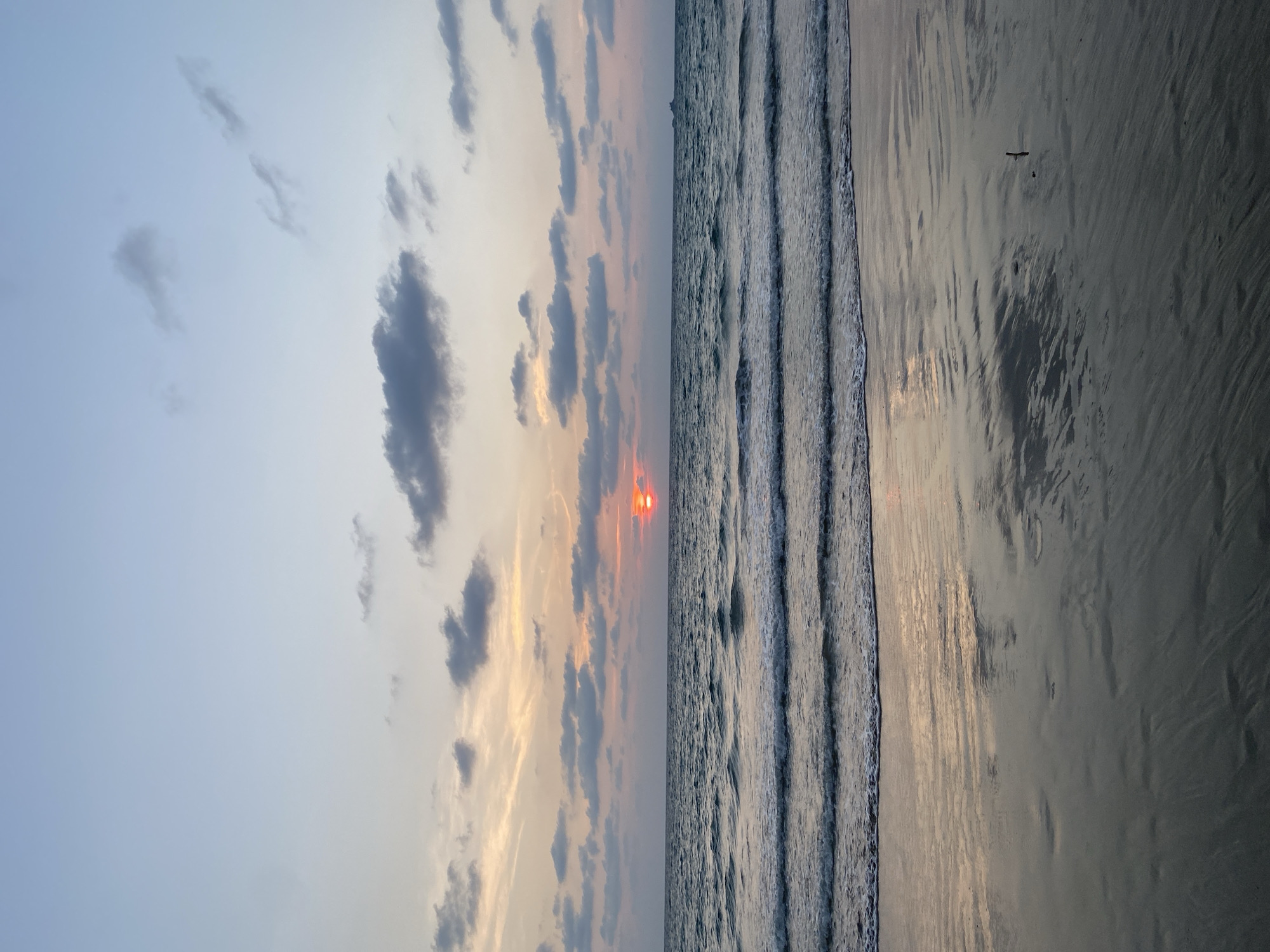 The image depicts a beach scene at sunset in Choeng Thale, Thailand. The sky features a mix of blue and orange hues with scattered clouds. The sun is visible as a red disk near the horizon. The ocean is characterized by visible waves moving towards the shore. The beach itself is smooth and wet, reflecting the sky and sunlight. A single bird is flying over the sand.