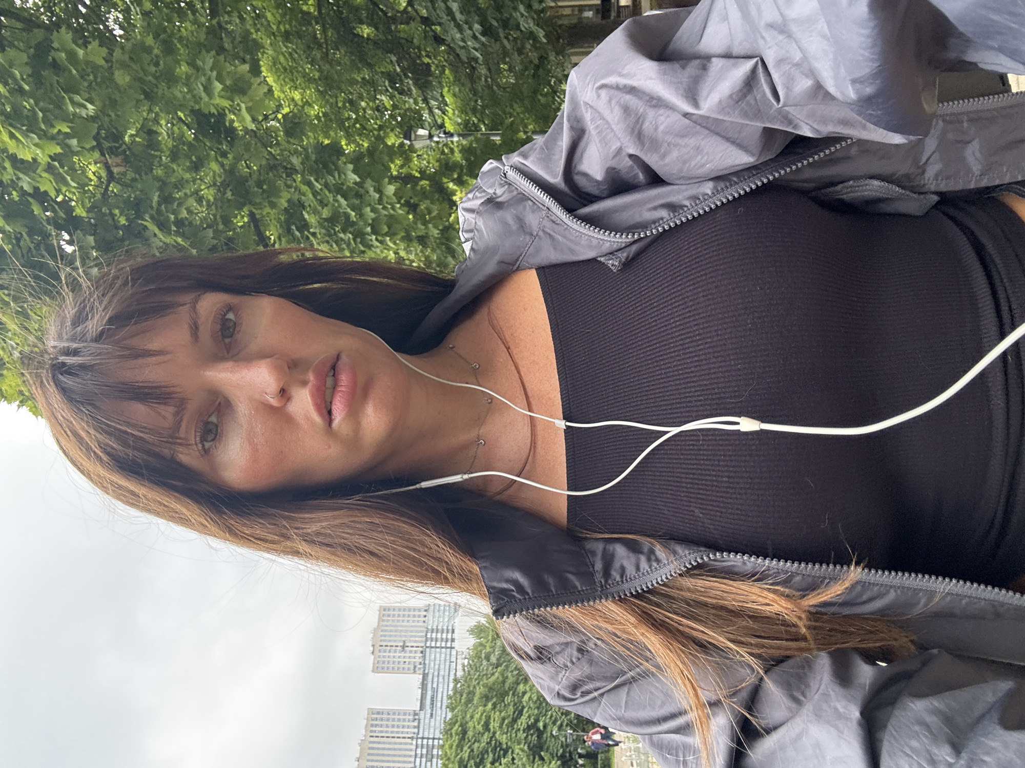 A selfie is taken by a woman with long brown hair and bangs, wearing a black ribbed tank top and a grey zippered jacket. She has a nose ring and is wearing white earbuds. The background shows green trees and a cloudy sky, with tall buildings visible in the distance, suggesting an urban setting. The overall scene appears to be a park or public space within a city, possibly Moscow, Russia.