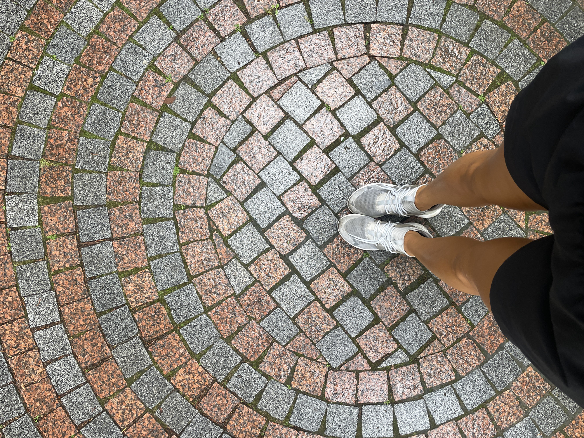 This is a top-down, eye-level shot of a person's legs and feet standing on a circular cobblestone pathway. The pathway is composed of alternating light grey and reddish-orange granite square stones. There are small patches of green moss or grass growing in the gaps between the stones. The person is wearing white athletic shoes with grey accents and black shorts. The lighting suggests an overcast day. The location is a public outdoor space in the city of Sochi, Russia.