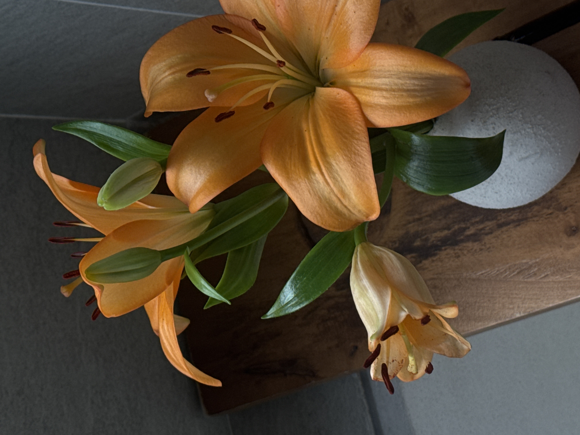 A close-up, high-angle shot captures a bouquet of orange lilies in a round, textured white vase. The lilies are in various stages of bloom, with some fully open and others still in bud. Their petals are a vibrant orange, with darker, reddish-brown anthers visible in the center of the open flowers. Green leaves and stems are interspersed among the blooms. The bouquet is set against a backdrop of a dark, textured wall and a wooden surface. The overall impression is one of natural beauty and simplicity, with the warm tones of the lilies providing a striking contrast to the cooler tones of the background. There are no people, animals, or discernible city elements present in this image.
