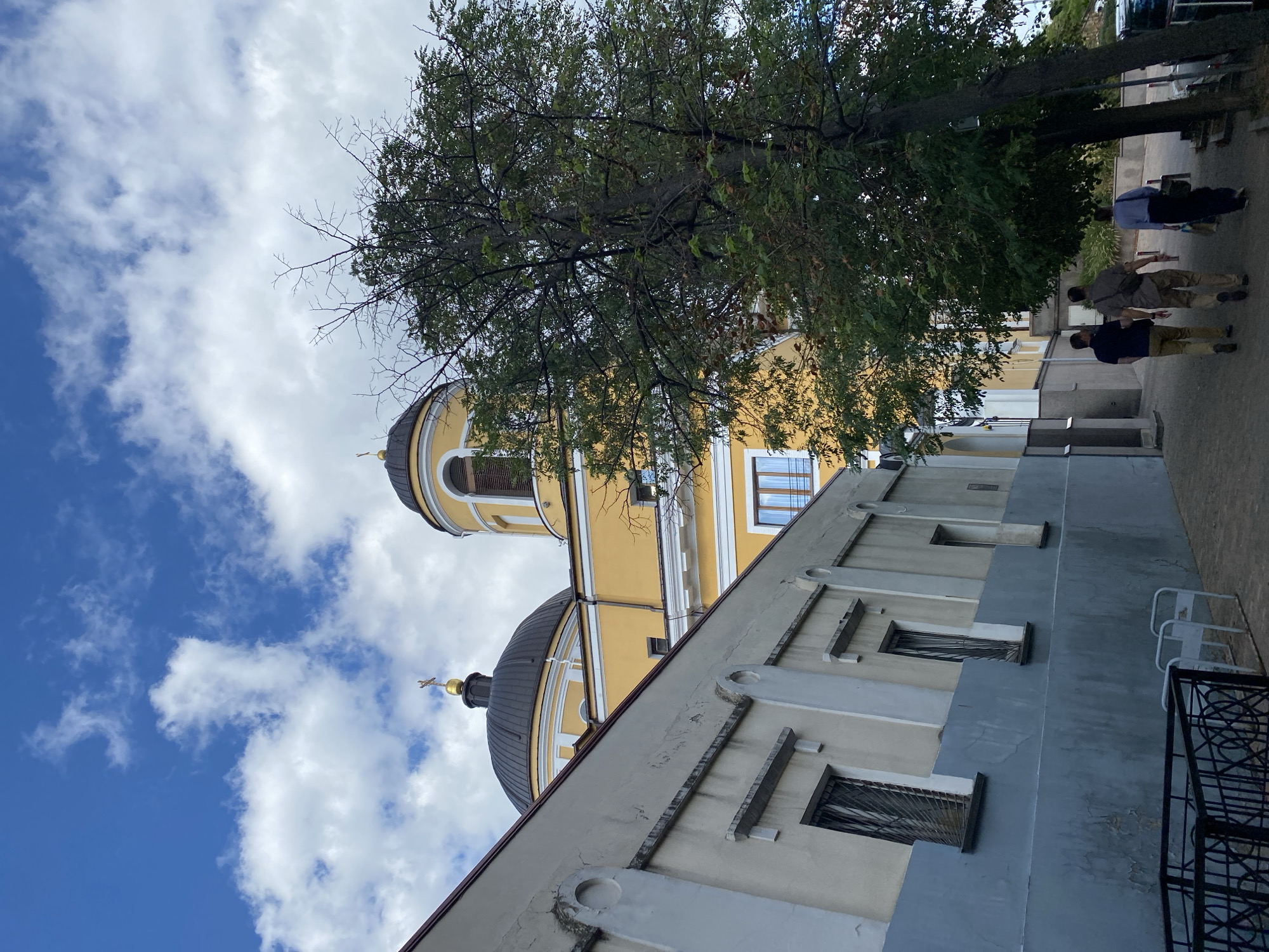The image captures a view of a yellow church building with a dark dome and a golden cross on top, situated in a city environment. In the foreground, a light-colored building runs along a paved walkway. A large tree with green foliage stands to the right of the church. Three individuals are walking away from the viewer on the walkway, with one carrying a bag. The sky is blue with scattered white clouds. The scene appears to be outdoors on a bright day.