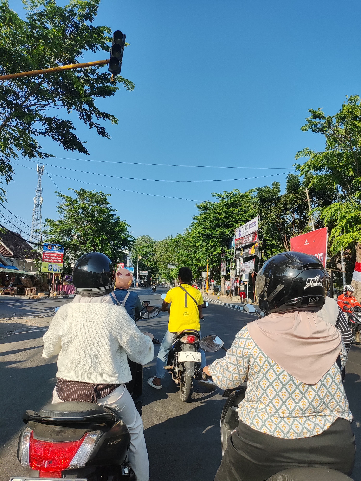 The image depicts a street scene in Cirebon, Indonesia, under a clear blue sky. Three motorcyclists are traveling on a road lined with trees and buildings. The first motorcyclist in the foreground is wearing a white knitted sweater and a black helmet. The second motorcyclist, seated behind the first, wears a light brown headscarf and a patterned long-sleeved shirt, also with a black helmet. The third motorcyclist, in front of the other two, is wearing a yellow t-shirt and blue jeans, with a black helmet. A fourth motorcyclist in an orange jacket is visible further ahead on the right. Billboards and traffic signals are present along the roadside, indicating an urban setting. The scene appears to be captured from the perspective of a passenger on one of the motorcycles.