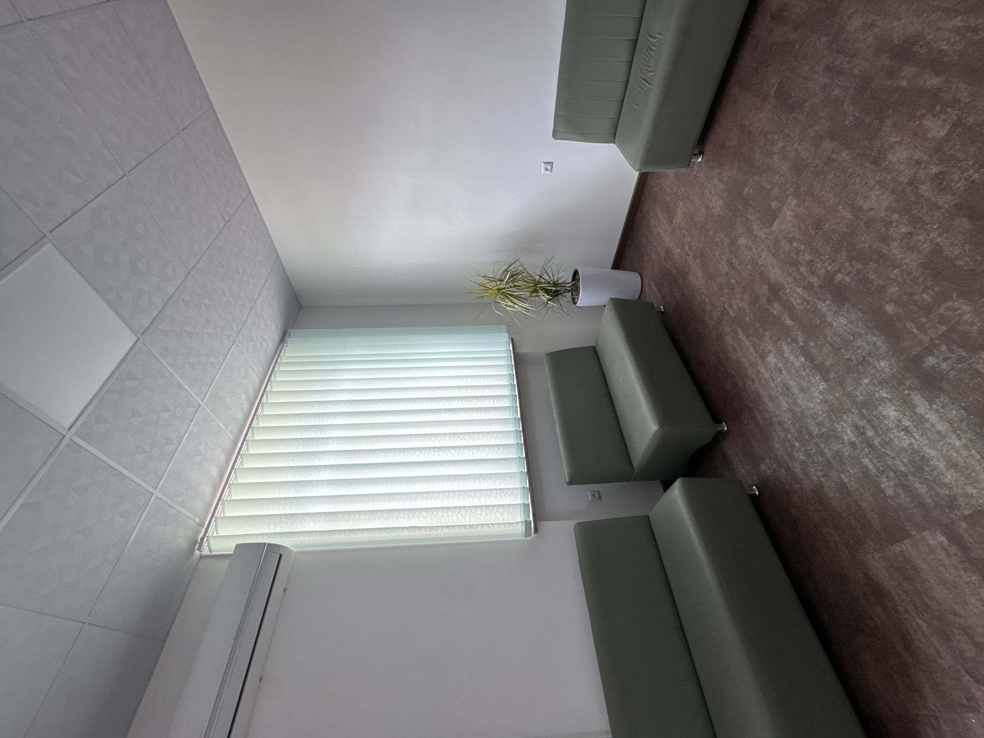 The image shows an interior space with seating arrangements. On the right side, there's a brown patterned floor, adjacent to which are two light green benches. A plant in a white pot sits near one of the benches. In the upper part of the image, there's another longer light green bench. A window with closed blinds is visible on the left side, and underneath it, an air conditioning unit is mounted. The walls appear to be white, and the ceiling has a patterned tile design. The scene seems to be a waiting area. There is no information about the location in the city of Yerevan, Armenia.