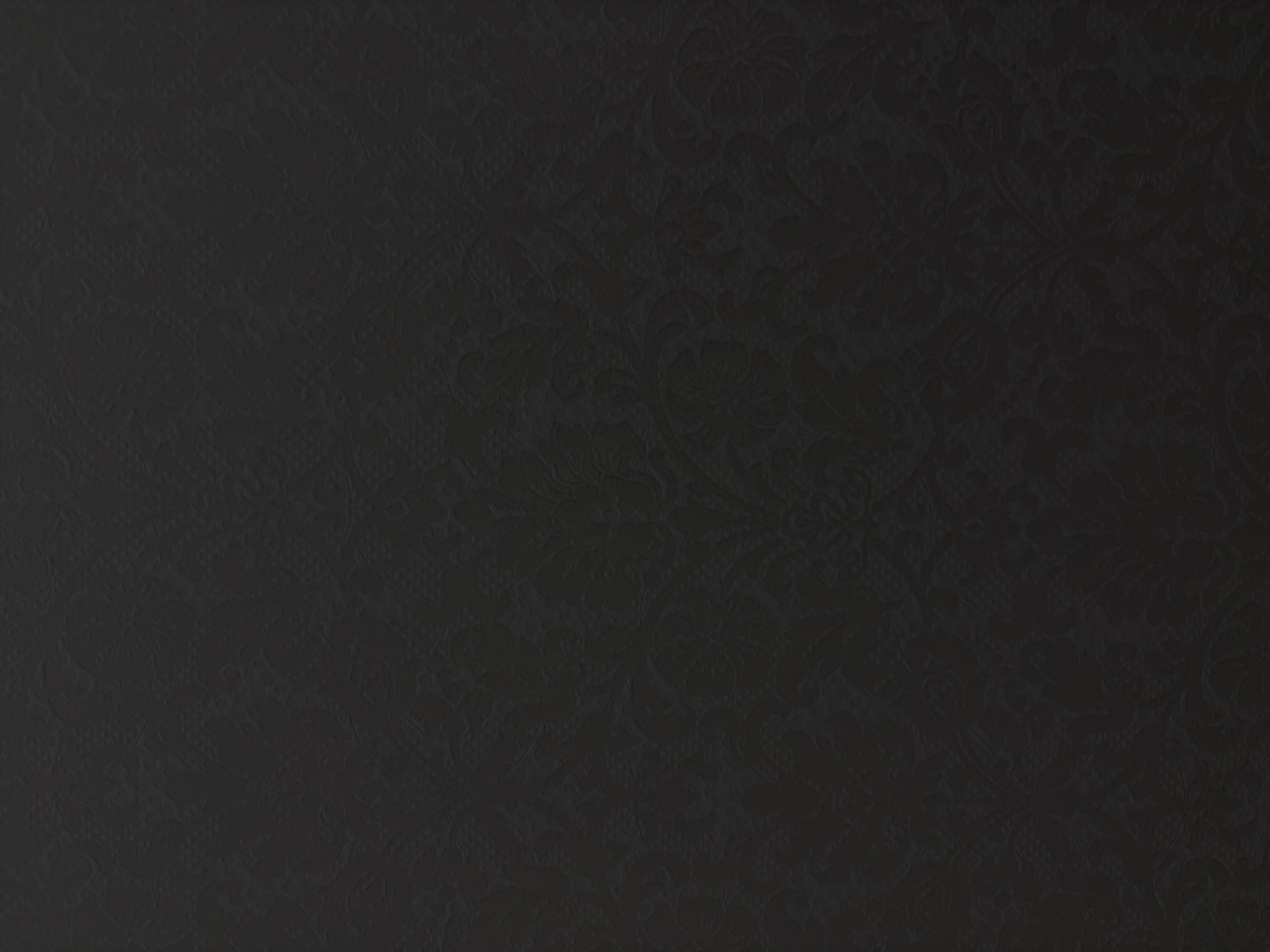 The image displays a dark charcoal grey to black surface, uniformly covered with a subtle, repeating embossed or debossed floral and damask pattern. The intricate design features swirling acanthus leaves, stylized blossoms, and scrollwork, discernible through minimal variations in light and shadow that define its low relief texture. The surface is evenly illuminated, highlighting its consistent dark tone and the delicate details of the pattern. No other subjects, objects, people, or actions are depicted. This textured surface is located within the city of Minsk, Belarus.