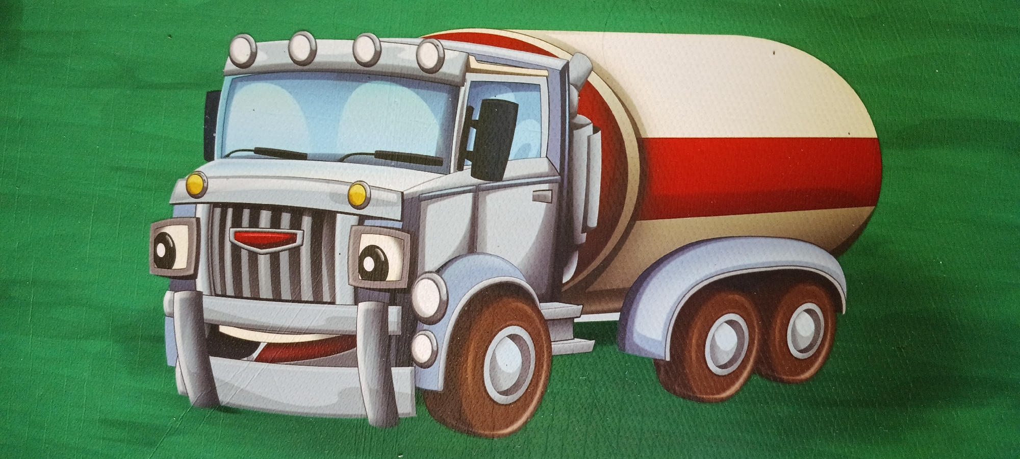 The image shows a cartoon illustration of a cement mixer truck. The truck is predominantly light blue and grey, with a white and red striped tank. It has large brown wheels, a smiling face with eyes, and headlights. The background is a textured green surface, possibly a wall or a mat. There are no people visible in the image, and no specific landmarks or context are provided to associate it with Denpasar, Indonesia.