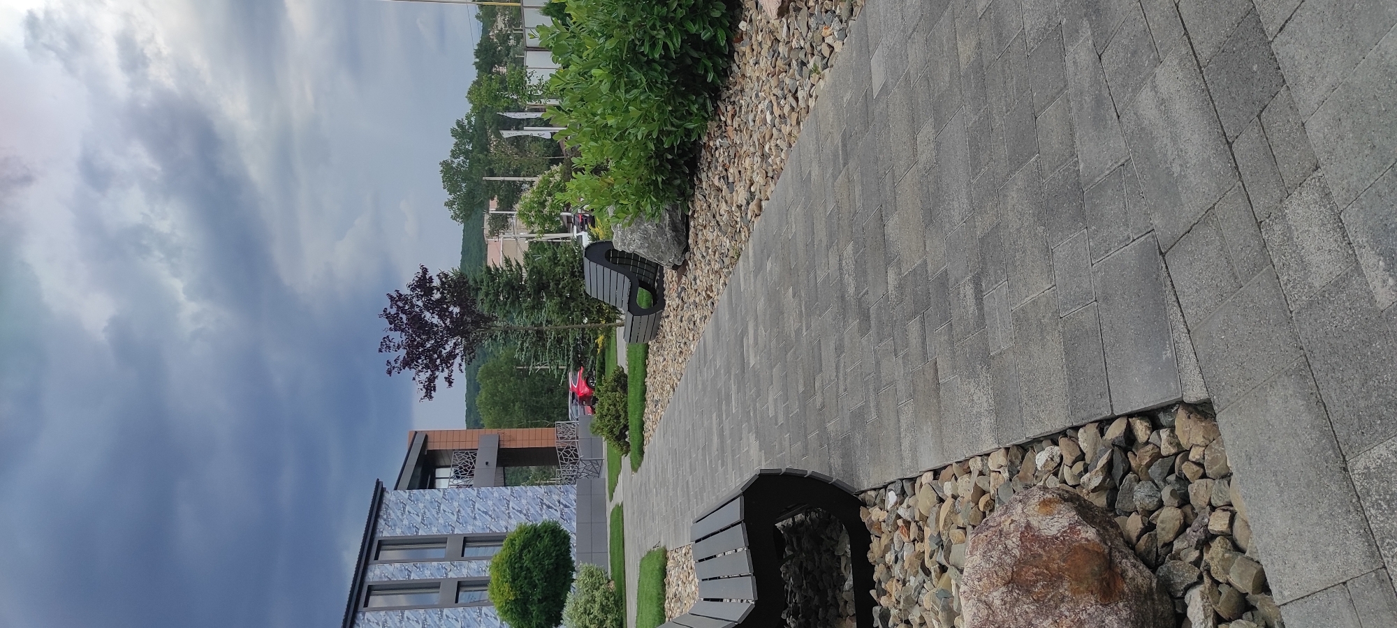 The image shows a modern building with a blue and grey facade, featuring large windows. In front of the building, there is a paved walkway made of grey interlocking bricks. Along the walkway, there are benches, decorative rocks, and manicured greenery. A dark red car is parked in the background. The sky is overcast with dramatic, dark clouds. The scene appears to be an outdoor public space or the grounds of a commercial or residential complex. There are no people visible in the image. The context of Tulskiy, Russia, is not visually evident from the image itself.
