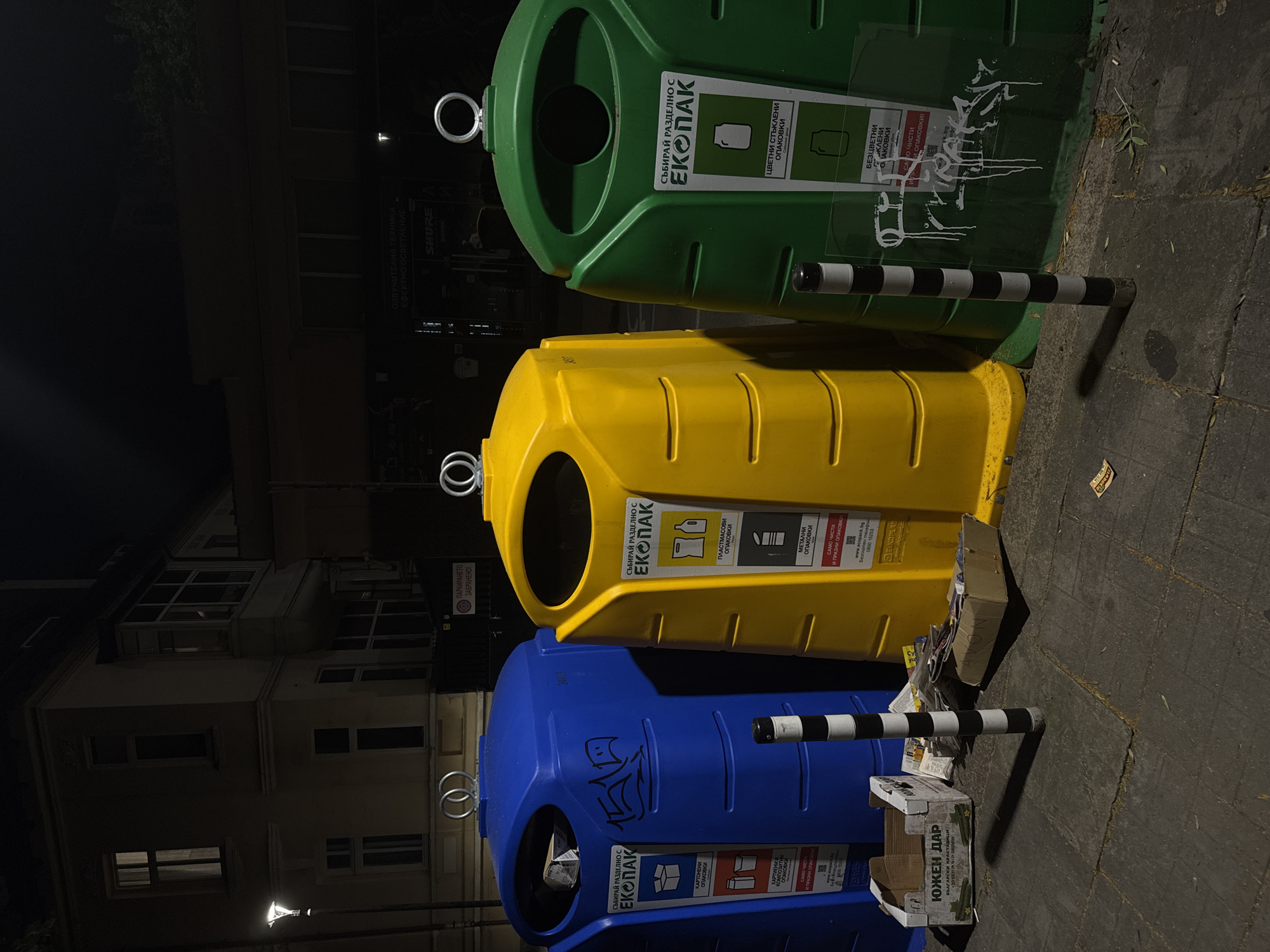 The image shows three large, colorful recycling bins (blue, yellow, and green) lined up on a sidewalk at night in Sofia, Bulgaria. The bins are labeled with "ECOPAK" and instructions in Bulgarian on what materials to deposit. Graffiti is visible on the blue bin. Various pieces of trash, including cardboard boxes and paper, are scattered on the sidewalk around the bins. In the background, a building with illuminated windows and a street lamp are visible. The scene suggests a typical urban environment at night with a focus on waste management infrastructure.