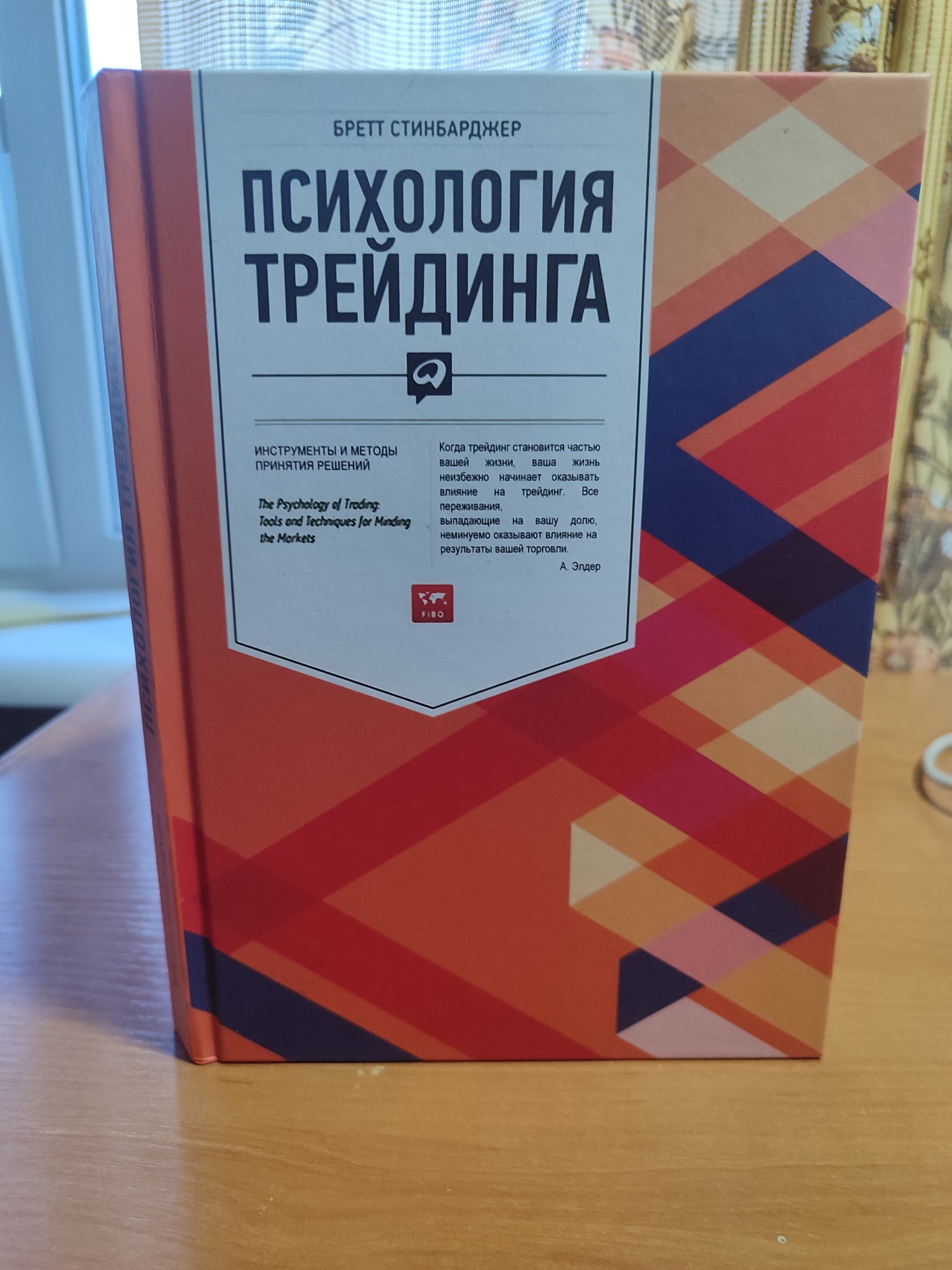 The image shows the cover of a book titled "ПСИХОЛОГИЯ ТРЕЙДИНГА" (Psychology of Trading) by Бретт Стинбарджер (Brett Steenbarger). The book cover features a geometric pattern in shades of orange, blue, and pink. Text on the cover includes the title, author's name, and a description of the book's content in Russian and English. Below the main text, there is a small logo that reads "FIBO". The book is placed on a wooden surface, likely a table, with a shadow cast on the left side, suggesting it is near a window or light source. The background is blurred, but appears to be a textured fabric, possibly a curtain or tablecloth. There are no people or animals visible in the image. The overall impression is that of a book cover presentation.