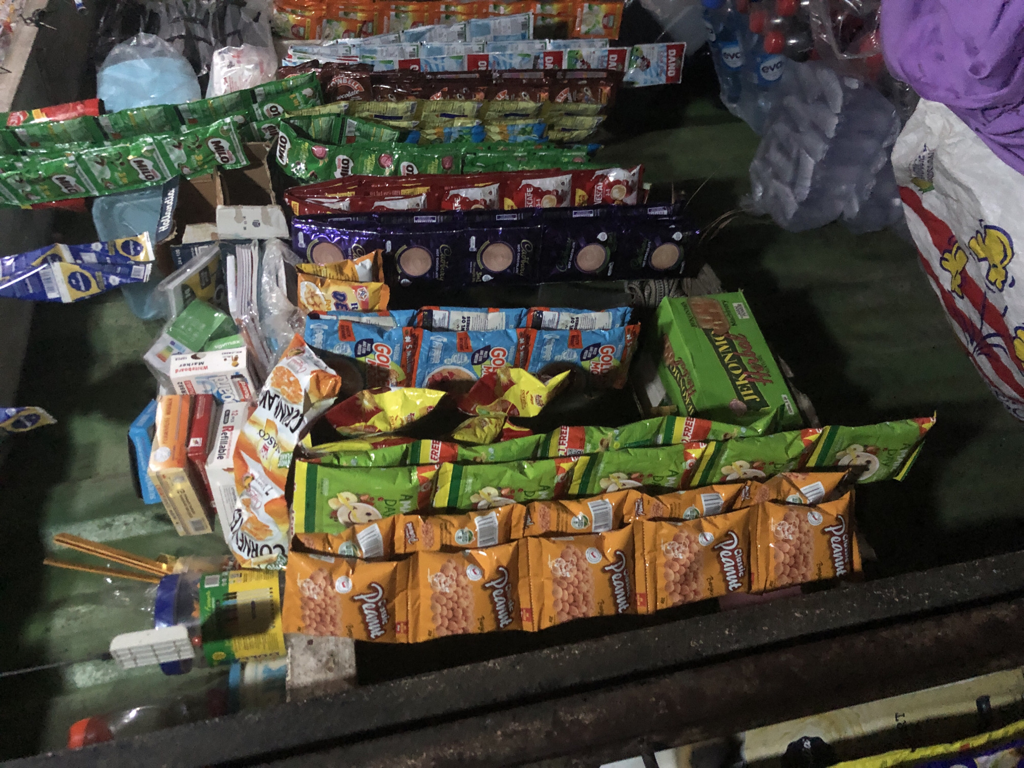 This is a medium shot, eye-level, indoor photograph of a small, cluttered roadside stall in Ado, Nigeria. The stall appears to be selling a variety of convenience items, including snacks, drinks, and household goods. The overall impression is one of abundance and perhaps a lack of organized display, typical of many informal businesses in the region.
In the foreground, a collection of individually wrapped snacks are hanging from a string or wire. These include several packages of "Peanut Classic" snacks, recognizable by their orange packaging and image of peanuts. Next to these are packets of green-colored snacks, some of which bear the text "FREE". To the right, there are more hanging packets of snacks and drinks, including some with the "Milo" logo and others with red and white packaging. Further to the right, several bottles of drinks are visible, some in plastic packaging.
Behind the hanging snacks, the stall's inventory is stacked. There are boxes of what appear to be crackers or biscuits, some in yellow and orange packaging, with visible text like "CORNE" and "NASC". There are also stacks of smaller items, possibly batteries or other small electrical components, and some packaged toiletries. In the background, a green box with "JEKONMC Herbal" written on it is visible.
The stall itself is constructed with rough materials, including corrugated metal sheeting on the left side, suggesting it's a temporary or semi-permanent structure. The lighting is dim, indicating an interior or shaded area, possibly illuminated by artificial light or natural light filtering in from an opening. The background is dark and indistinct, providing little information about the broader environment. There are no people visible in the frame. The overall scene suggests a busy, working stall, likely catering to the local community.