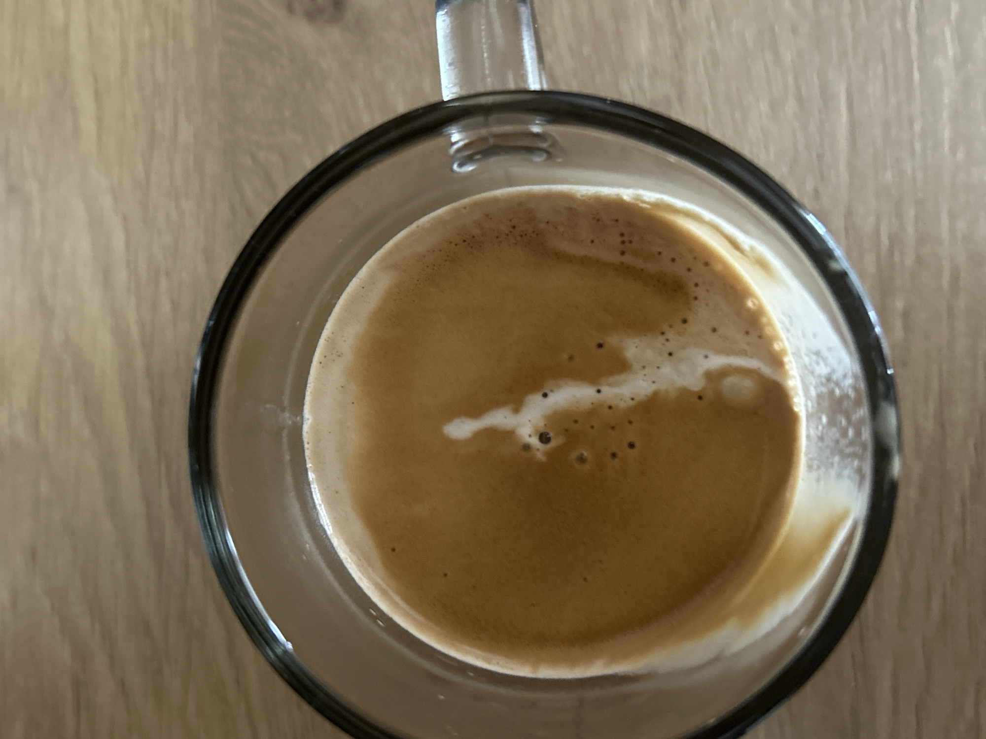 A top-down view shows a clear glass mug containing coffee, possibly an espresso. The coffee has a light brown color with some crema and small white froth details on the surface. The mug has a handle and sits on a wooden surface. There are no people visible. The location in the city of Sofia, Bulgaria, cannot be determined from the image.