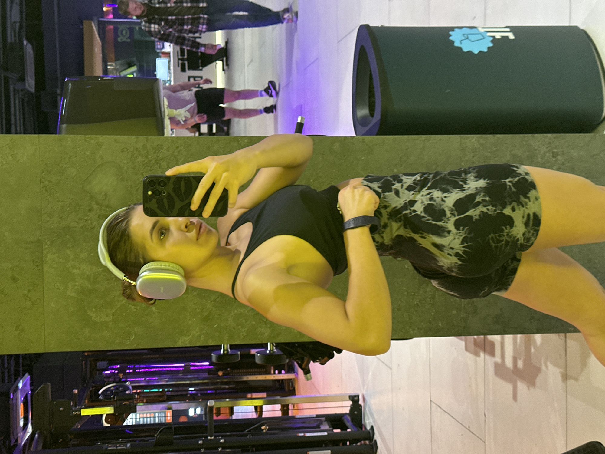 The image depicts a woman taking a selfie in what appears to be a gym setting. She is wearing headphones, a black tank top, and patterned shorts. The gym equipment is visible in the lower part of the image.
The location is in Irkutsk, Russia, based on the presence of a water cooler branded with the "Baikal Water" logo, a company located in the area. There are people in the background on a different floor.