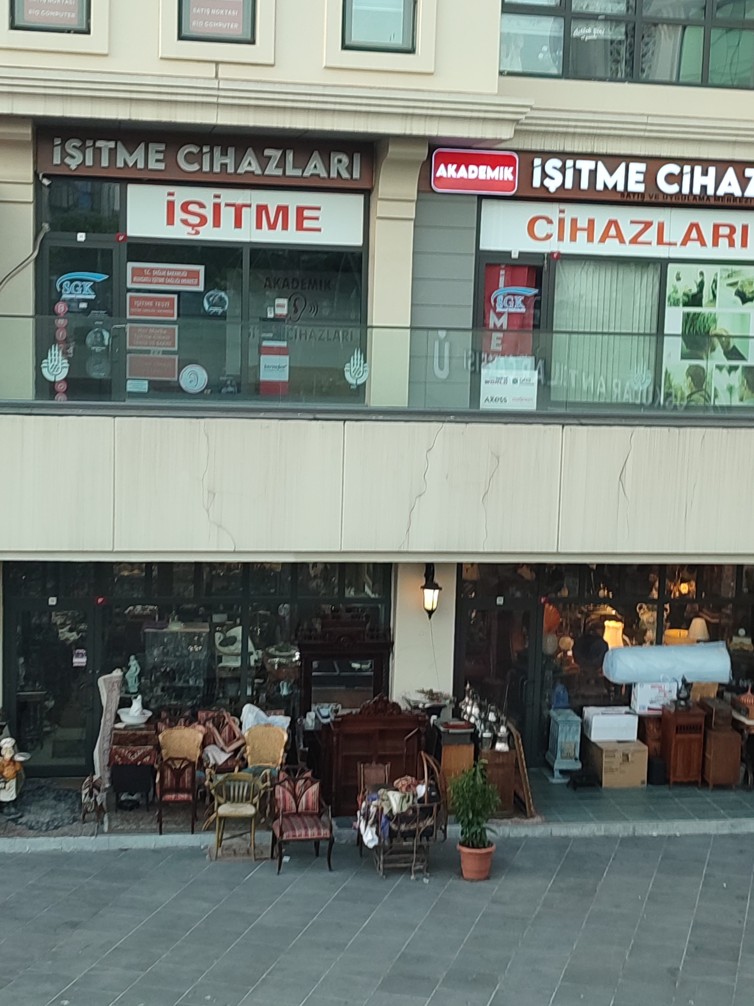 The image shows the storefronts of businesses located in Üsküdar, Turkey, likely on a commercial street or in a shopping center. The businesses appear to be related to hearing aids and possibly antique or vintage items.
In the upper section, there are two prominent signs for businesses. The sign on the left reads "İŞİTME CİHAZLARI" (Hearing Aids) with a smaller sign below it saying "İŞİTME" (Hearing). The sign on the right also reads "İŞİTME CİHAZLARI" and has the word "AKADEMİK" (Academic) in a red oval above it, with "CİHAZLARI" (Devices) also visible. Through the glass windows of these businesses, various items and displays can be seen, including logos, informational signs, and what appear to be hearing aid devices or related products. One window has a sign that reads "AKADEMİK" next to an icon of an ear.
Below this upper level, there is a second row of storefronts. The windows of these shops reveal a collection of antique or vintage furniture and decorative items. Visible items include chairs of various styles, wooden cabinets, lamps, and other assorted objects. A small potted plant is placed outside one of these shops. The overall environment appears to be a pedestrian area or sidewalk in front of the shops. The lighting suggests it is daytime, possibly late afternoon, given the shadows. The architecture of the buildings is modern, with beige-colored panels and large glass facades for the storefronts.