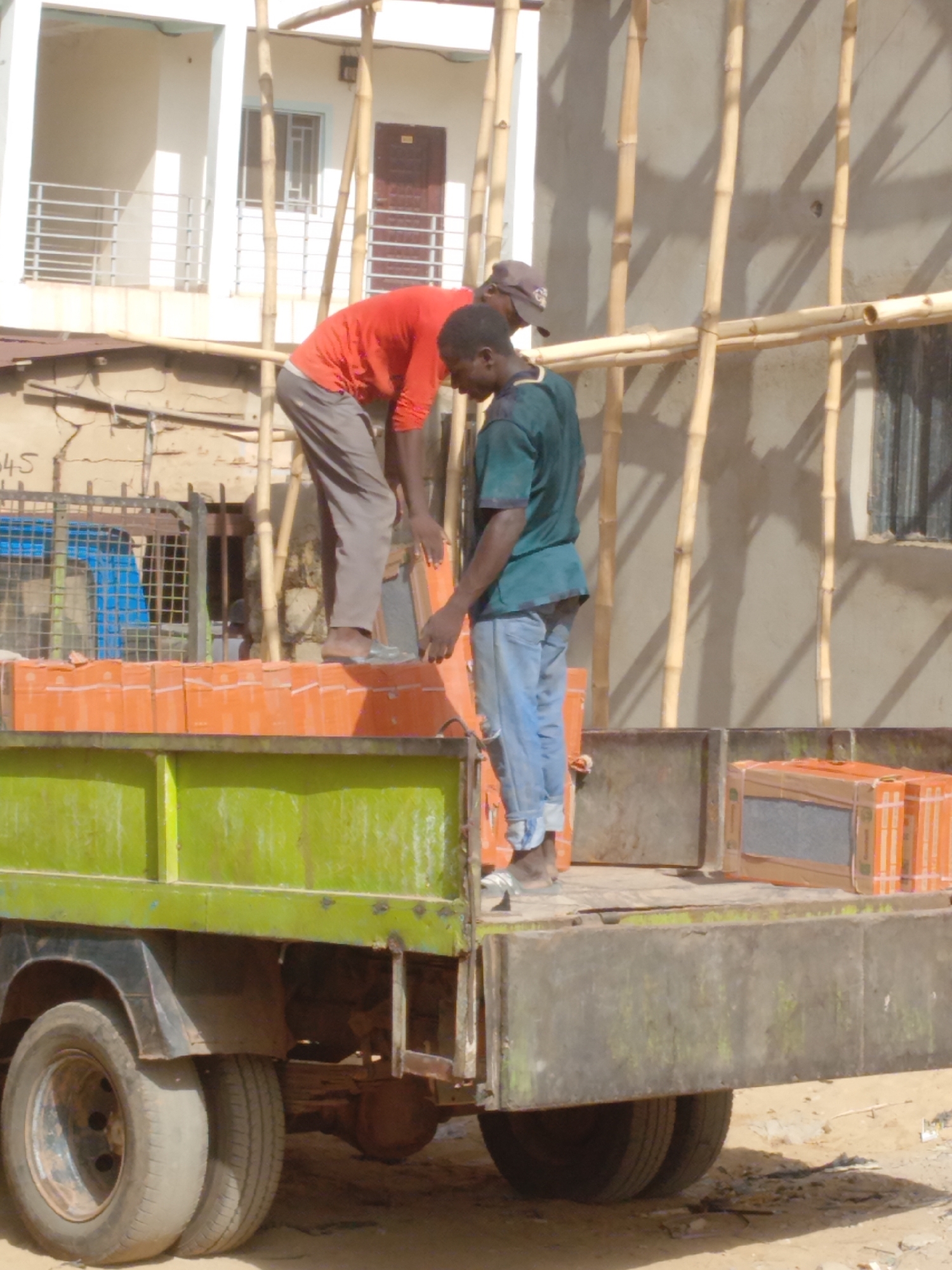 Two men are on the back of a green and grey truck, seemingly unloading orange-boxed materials. One man, wearing an orange shirt and grey pants, is bent over handling the boxed materials, while the other man, in a green shirt and blue jeans, is standing upright, also handling the boxed materials. The truck is loaded with more of these orange-boxed materials. In the background, there is a building under construction with bamboo scaffolding and a partially visible white building. The location appears to be the city of Sardauna, Nigeria. The scene suggests manual labor involved in unloading construction or building materials.
