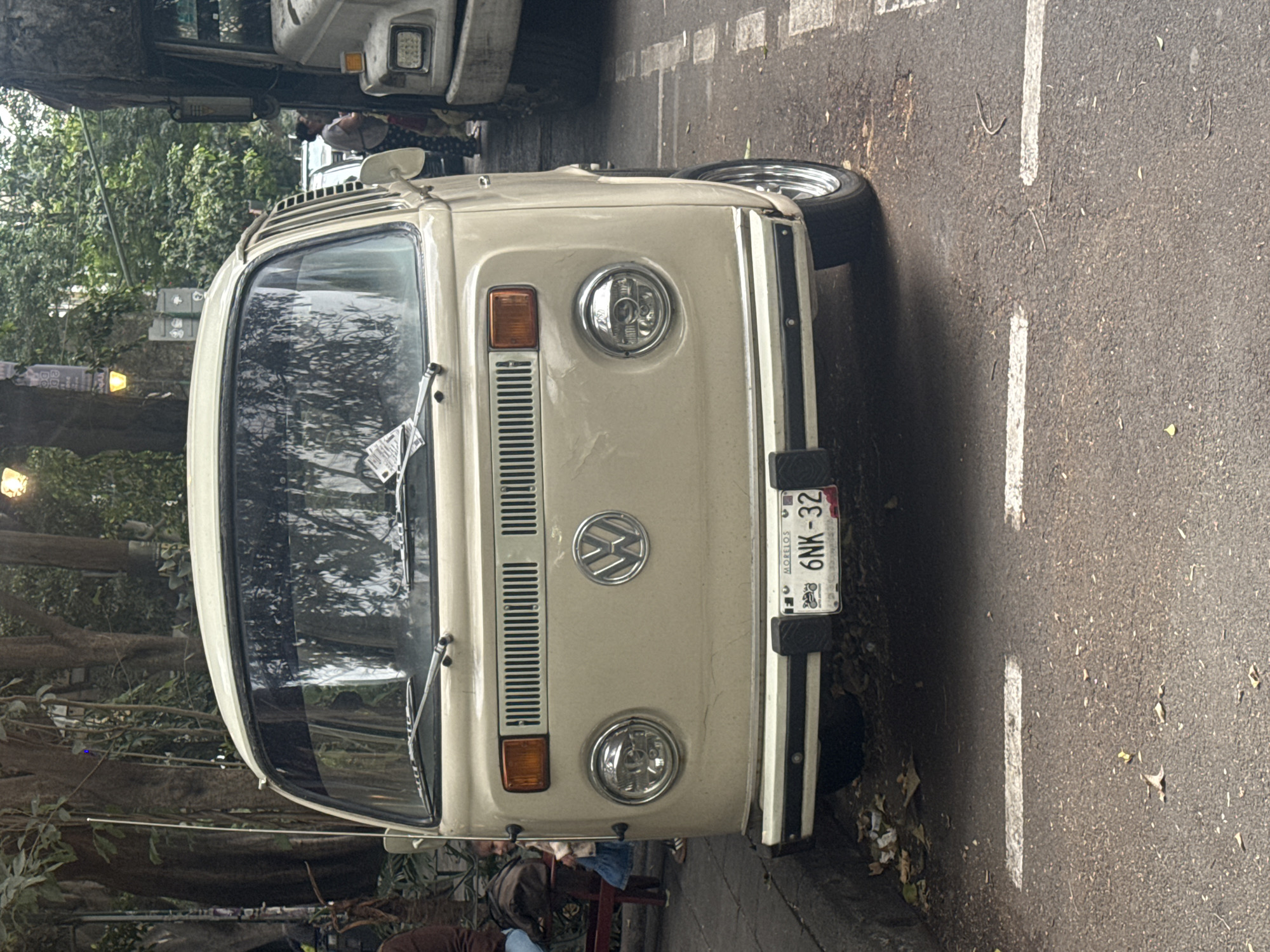 The image shows the front of a beige Volkswagen bus with the license plate "6NK-32" from Morelos, Mexico. The bus is parked on the side of a street with white painted lines. Trees and other vehicles are visible in the background, suggesting an urban environment, likely in Mexico City. The lighting is somewhat subdued. People are partially visible near the front of the vehicle.