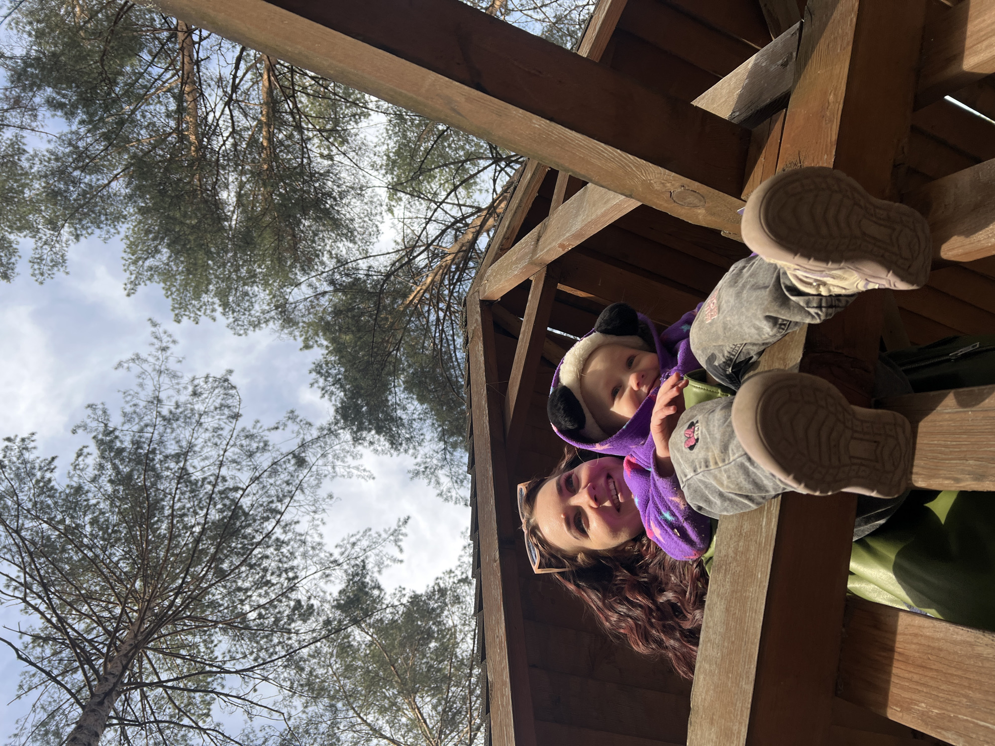 The photograph captures a scene with a woman and a young child. The woman has long, curly brown hair and is wearing sunglasses. She is smiling. The child is wearing a purple, hooded outfit with animal ears and a light-colored lining. The child's feet are visible with grey denim pants and boots.
They are located within a wooden structure, possibly a treehouse or similar construction, given the visible wooden beams. Through the beams, trees and a partly cloudy sky are visible.
The location may be Chaikovsky, Russia, but the photograph doesn't have any other indicators.