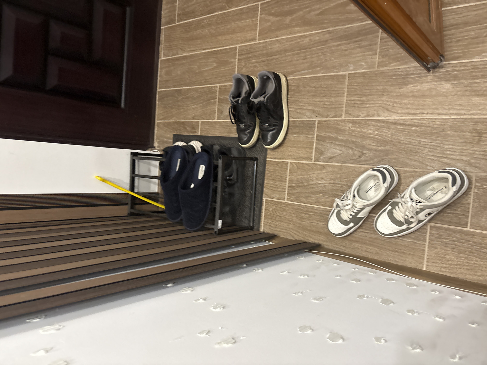 The image depicts a cluttered entrance area. On a tiled wall, a pair of black leather shoes is hanging above a metal shoe rack holding two navy blue slippers. Below, on the floor, rests a pair of white sneakers with grey accents. The floor is covered with a material that appears to have water spots. A brown door is visible on the left side of the image. The environment is indoor. No people are visible. The location is not discernible as Lovech, Bulgaria, based on the given image.