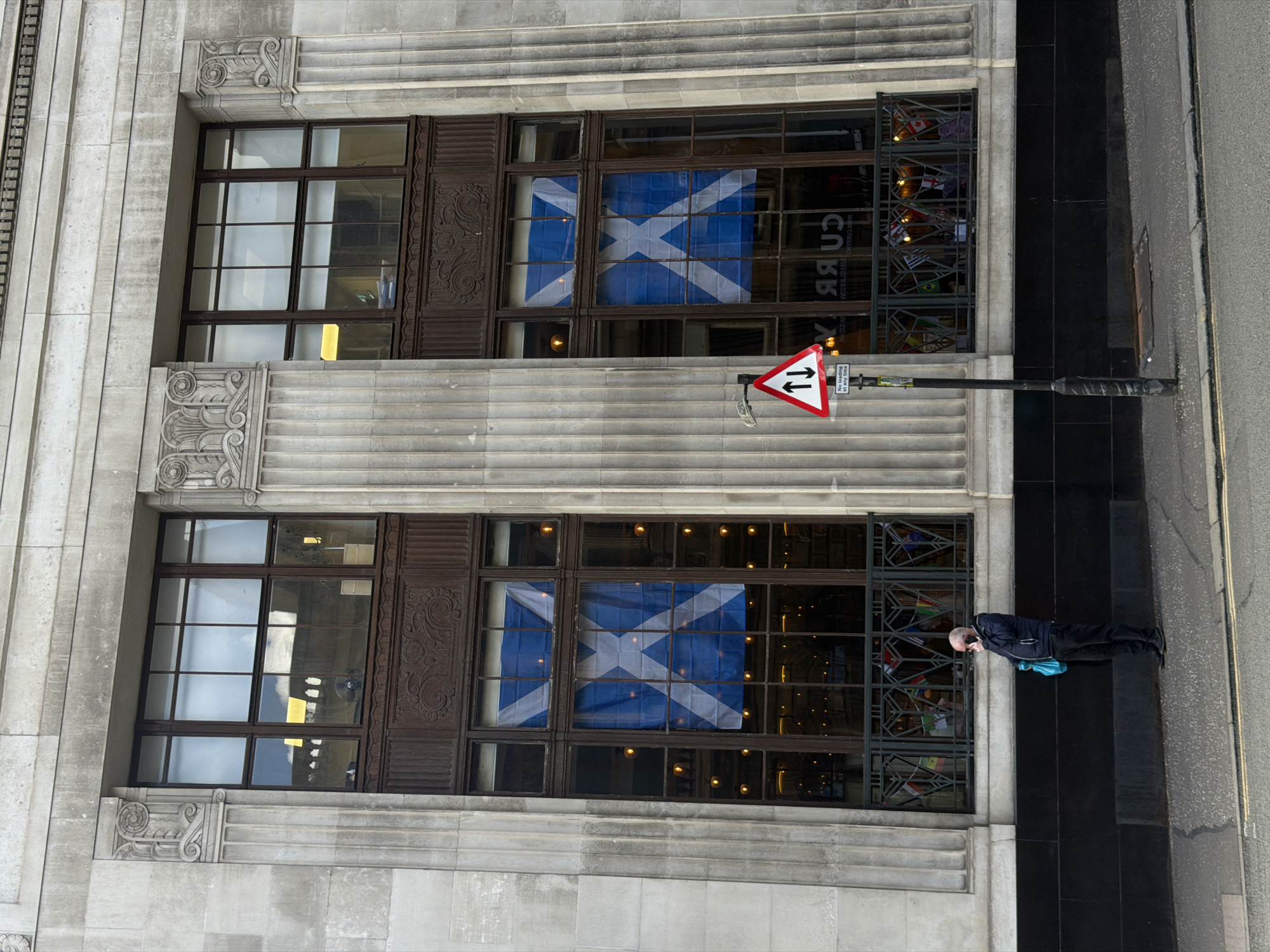 The image shows the facade of a stone building with large, ornate windows. The building is located in the city of Glasgow, United Kingdom. Two of the large windows on the second floor display Scottish flags. A person is standing on the sidewalk in front of the building, holding a light blue bag and a mobile phone to their ear. They are wearing dark clothing. A traffic sign is visible on a pole to the right of the person, indicating two-way traffic. The ground in front of the building is paved with asphalt and concrete. The facade of the building is made of light-colored stone with decorative elements and columns.