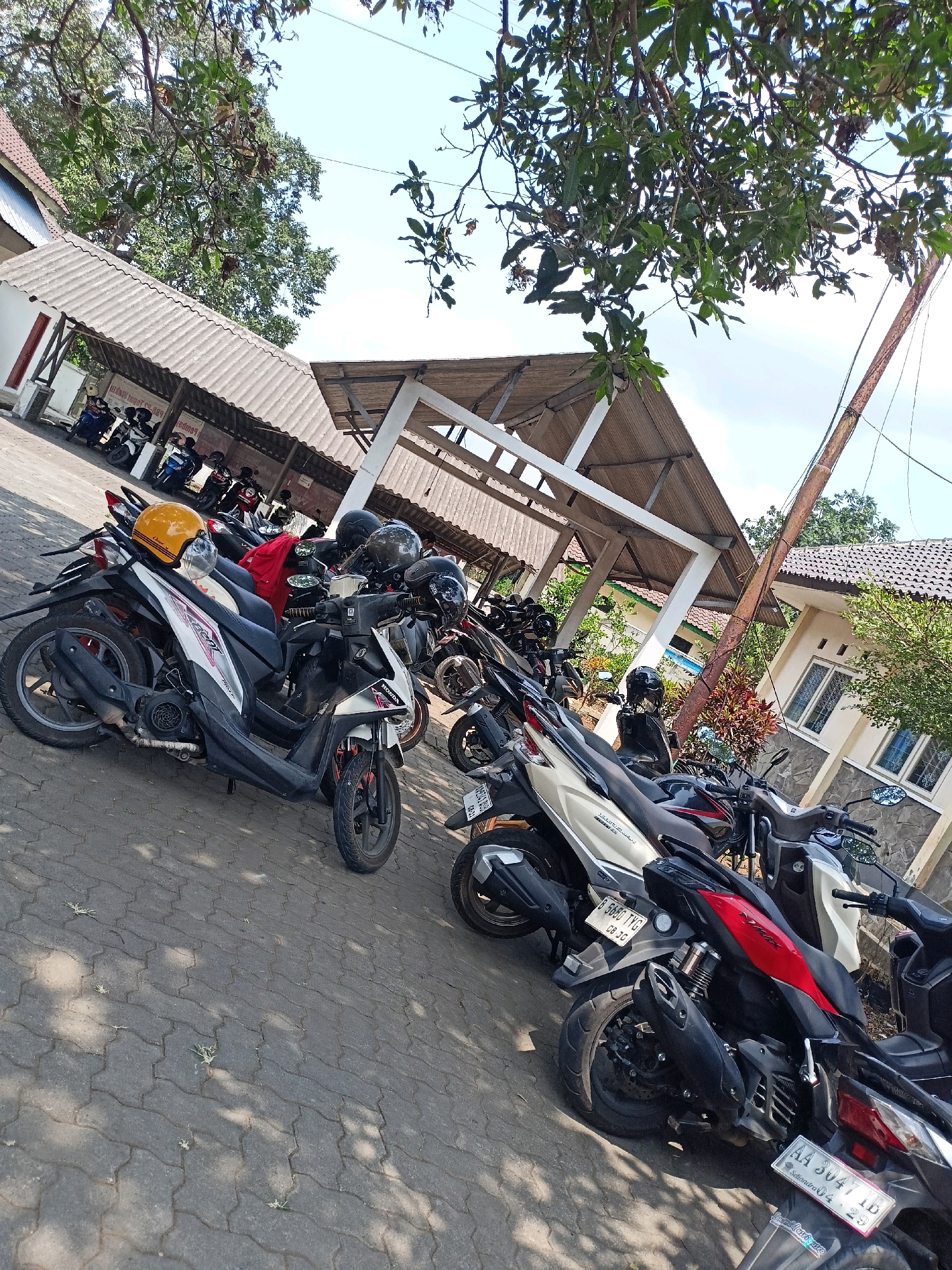 In the city of Kewaluhan, Indonesia, a paved outdoor area adjacent to a building with a tiled roof and open-air shelter is filled with parked motorbikes. The motorbikes, predominantly scooters, are lined up in rows. A yellow helmet is placed on the seat of a white and black scooter in the foreground. Some motorbikes have helmets resting on their seats or handlebars. The ground is made of interlocking concrete pavers, with patches of grass growing through the gaps. Sunlight casts shadows from the overhead trees and the shelter onto the ground. The building behind the shelter appears to be a single-story structure with windows and a stone-like base. The sky is visible through the branches of trees.