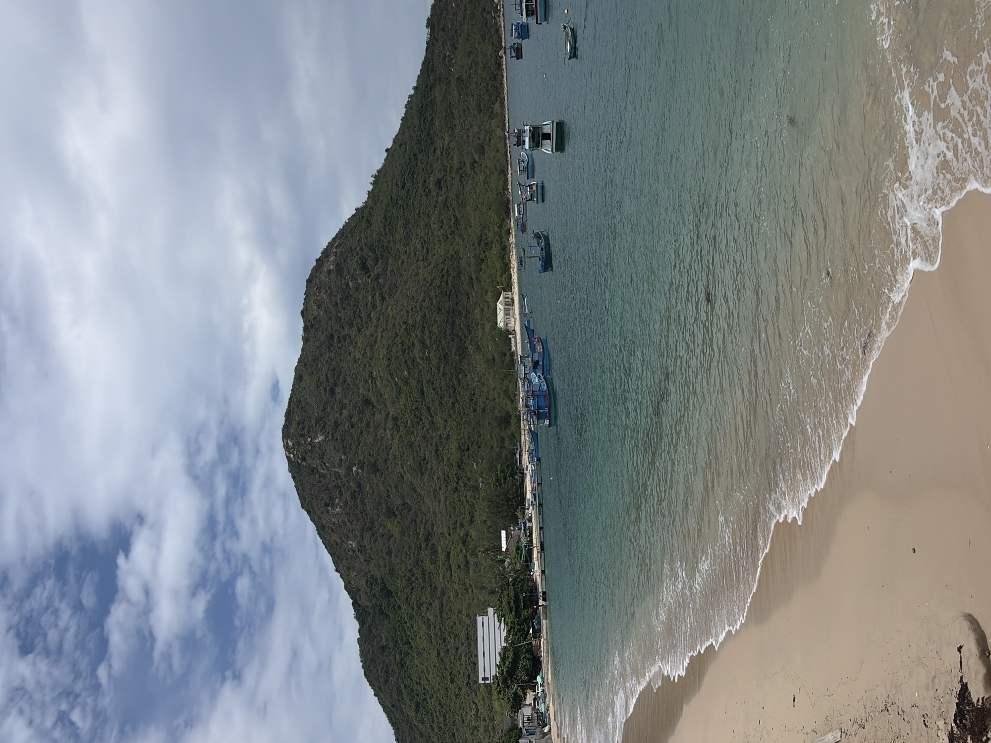 The image depicts a serene coastal landscape in Đông Ninh Hòa, Vietnam. On the left, a majestic, steep mountain covered in lush green foliage dominates the scene, extending its base down to the water's edge. Above the mountain, a bright sky with scattered white and grey clouds is visible.
In the center, a calm body of water, likely a bay or sea, stretches across the frame, displaying various shades of green and blue. Numerous small to medium-sized boats, appearing to be fishing vessels, are anchored or docked in the water, particularly along the base of the mountain. Several modest buildings and structures are discernible along the narrow strip of land between the mountain and the water, suggesting a small coastal settlement or harbor.
To the right, a sandy beach curves along the shoreline, meeting the gentle waves that break softly, forming foamy white lines. The sand appears light brown. No people are clearly visible, and the boats appear stationary, suggesting a tranquil moment in a working coastal environment. There is no discernible text in the image. The scene captures the peaceful coexistence of nature and human activity in a coastal Vietnamese setting.
