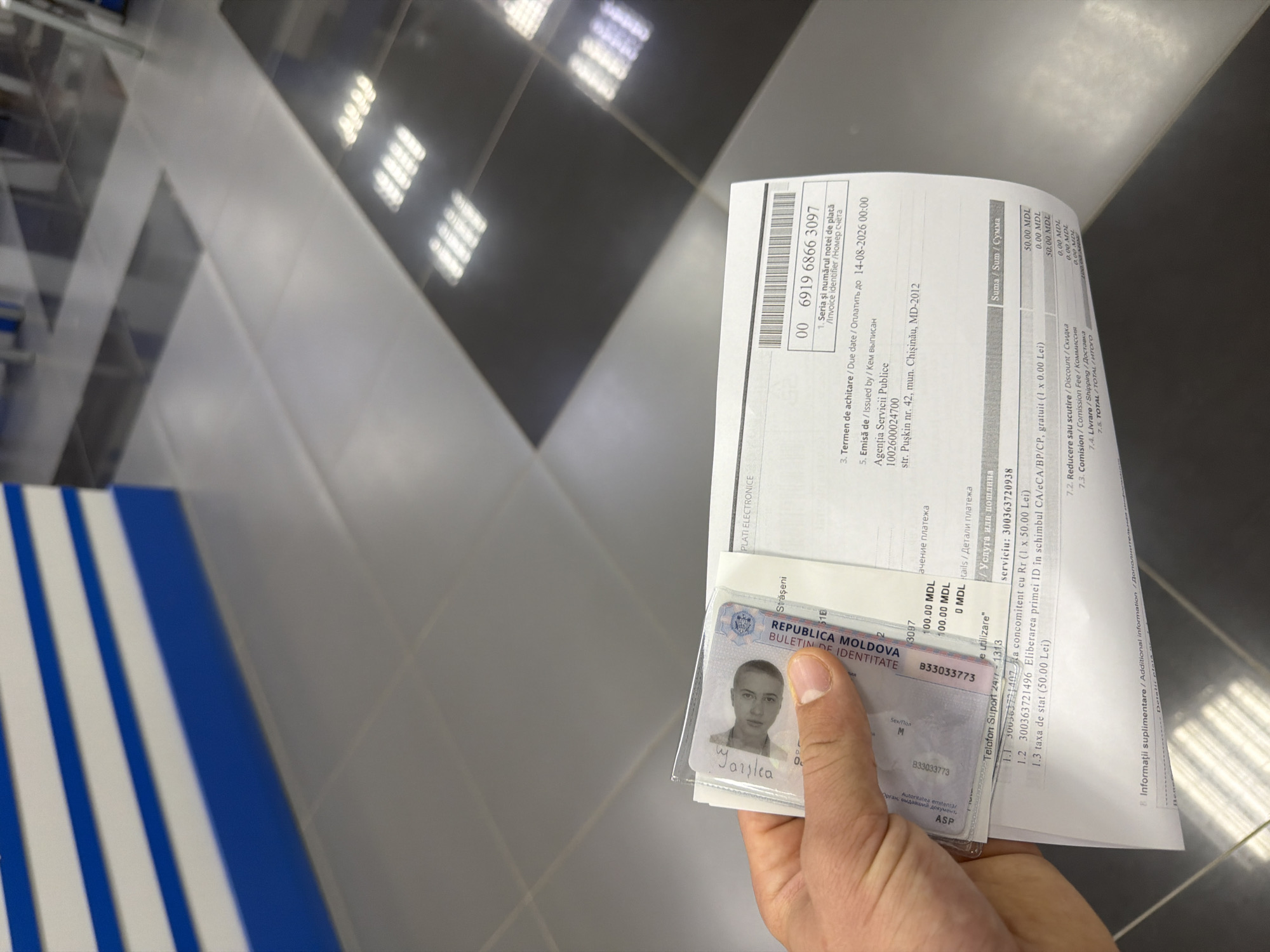 A person's hand is holding up a Moldovan ID card and a receipt. The ID card features a portrait of a person and text including "REPUBLICA MOLDOVA" and "BULETIN DE IDENTITATE." The receipt shows various fields with text in Romanian and English, including "PLATI ELECTRONICE," "Seria și numărul notei de plată," and "Termen de achitare." It also displays a barcode and a serial number. Below the ID and receipt, the environment appears to be an indoor space with tiled flooring and a ceiling with fluorescent lighting. The bottom part of the receipt indicates "str. Pușkin nr. 42, mun. Chișinău, MD-2012," which is an address in Chișinău, Moldova, not Strășeni.