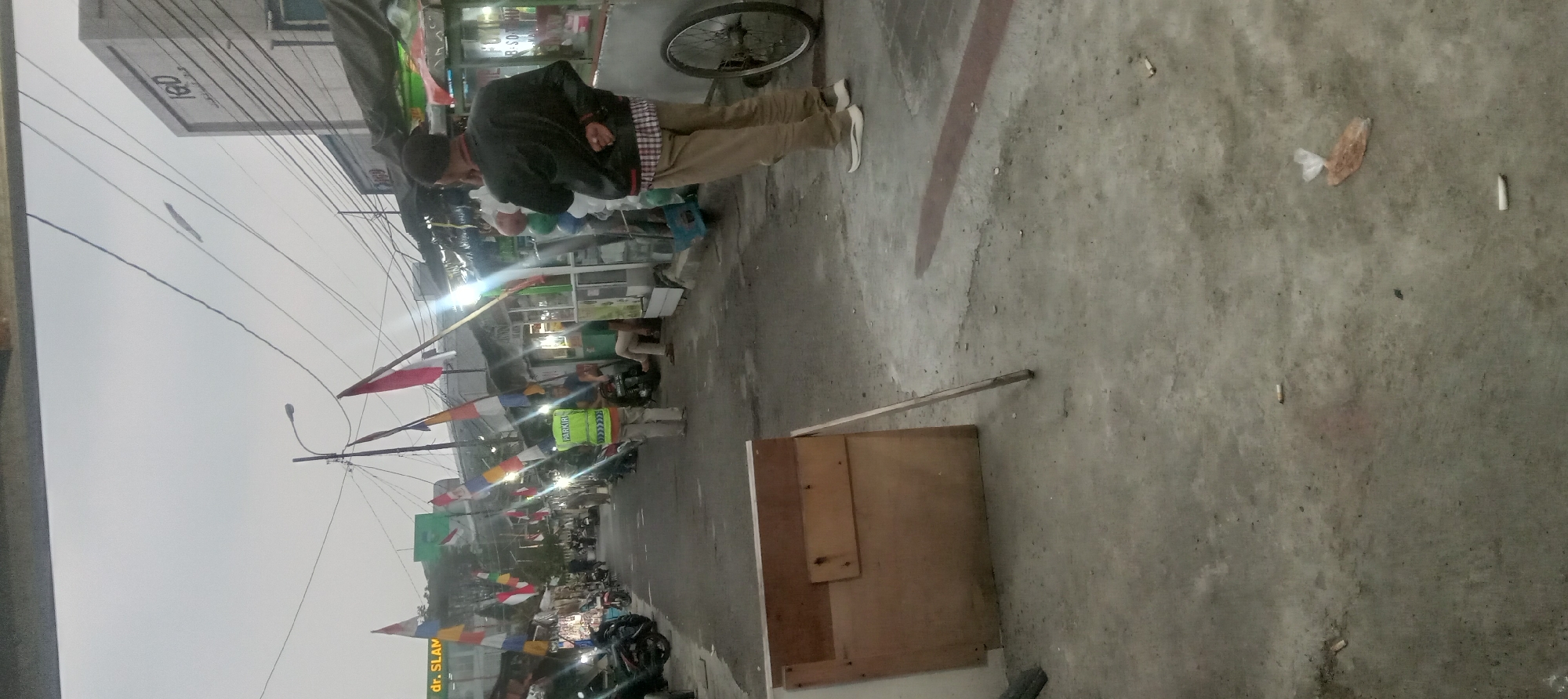 The image depicts a street scene in Garut, Indonesia. A man wearing a black jacket over a plaid shirt and tan pants stands with his back to the camera, talking on his phone. In front of him, a person in a bright yellow vest with "PARKIR" written on it stands facing the camera. The street is lined with various food stalls and shops. Red and white flags are displayed along the street. Several motorcycles are parked on the side. The ground is paved with concrete and shows signs of wear and tear, with scattered cigarette butts and a plastic bag. The overall atmosphere suggests a bustling, somewhat informal market area.