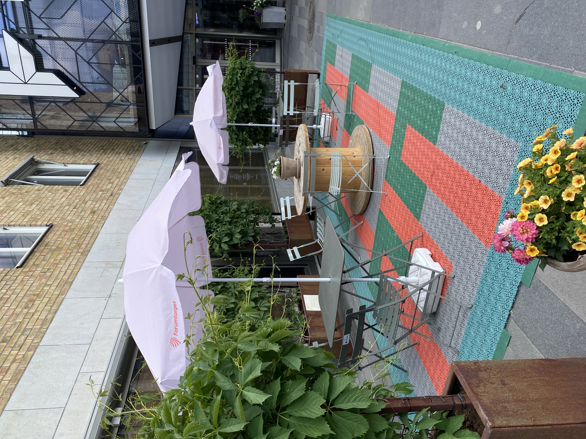 An outdoor seating area, likely a cafe or restaurant terrace, is viewed from an elevated perspective looking down. The scene is set in Uppsala, Sweden, on what appears to be a bright, possibly overcast, day. No people are visible.
The seating area features a mix of furniture, including several tables with light-colored metal chairs. Notably, two large wooden cable spools are repurposed as tables, each with a potted plant on top. Two prominent light pink umbrellas, partially open, provide shade. Both umbrellas display the text "Forumtorget" along with a logo resembling a stylized 'F' or wave pattern.
The ground is covered with distinctive patterned tiles arranged in a mosaic of colors, primarily orange, green, blue, and grey, creating a geometric design. Lush green foliage, including climbing vines, frames parts of the seating area, and potted plants with vibrant yellow and pink flowers add splashes of color.
In the background, a modern building with a complex glass and metal facade featuring geometric patterns is visible. To the left, a traditional brick building with windows can be seen. A dark grey paved area borders the patterned flooring, suggesting a sidewalk or public square. The overall impression is an inviting urban outdoor space.