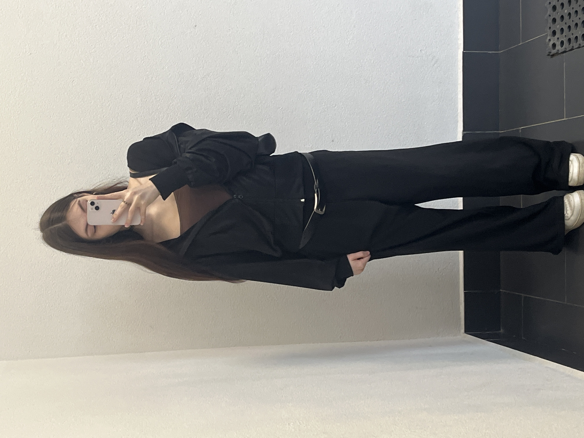 A female is taking a selfie in what appears to be an indoor hallway space. The subject, with long brown hair, is positioned at an angle. The person is wearing a brown top under a black jacket and matching black pants with a thin belt. She holds a phone, presumably to capture her reflection. The background features a light-colored wall with a dark tile area in the upper-right corner. The image does not allow inferring any information related to the city of Saint Petersburg, Russia.
