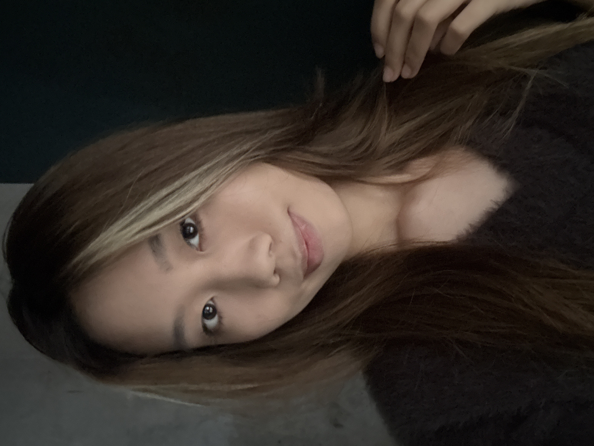 A close-up, angled portrait of a young woman with long, brown hair and a blonde streak near her face. She has brown eyes, fair skin, and appears to be wearing a dark, fuzzy sweater. Her right hand is partially visible, touching her hair near the top of the frame. The background is dark, likely a wall or dimly lit space. The photograph's location in Hechuan, China is not discernible from the image.
