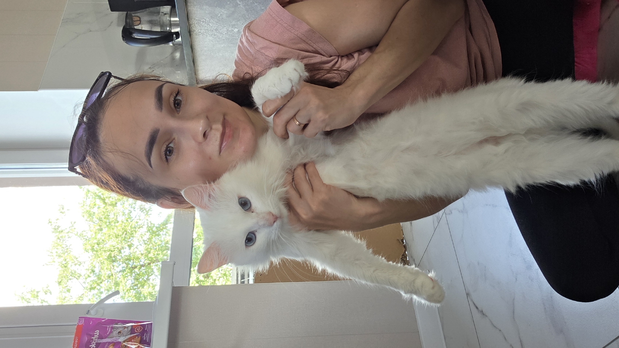 A woman is holding a white cat in a room that appears to be a kitchen. The woman has brown hair and is wearing a pink t-shirt and black pants. The cat is white with blue eyes. There is a window in the background with trees visible outside. A kettle and other kitchen appliances are on a counter behind the woman. The floor is tiled.