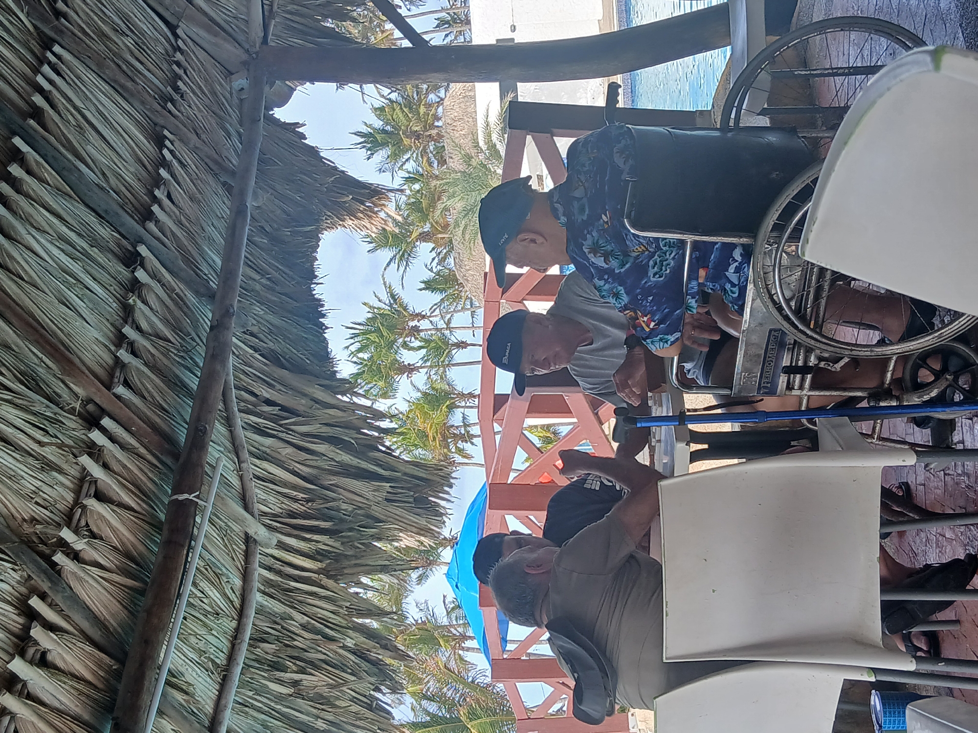 The image depicts a group of people seated at tables under a thatched roof structure in Altagracia, Venezuela. Several individuals are wearing caps, and one person is seated in a wheelchair. Palm trees are visible in the background, along with what appears to be a swimming pool. The scene is situated outdoors.