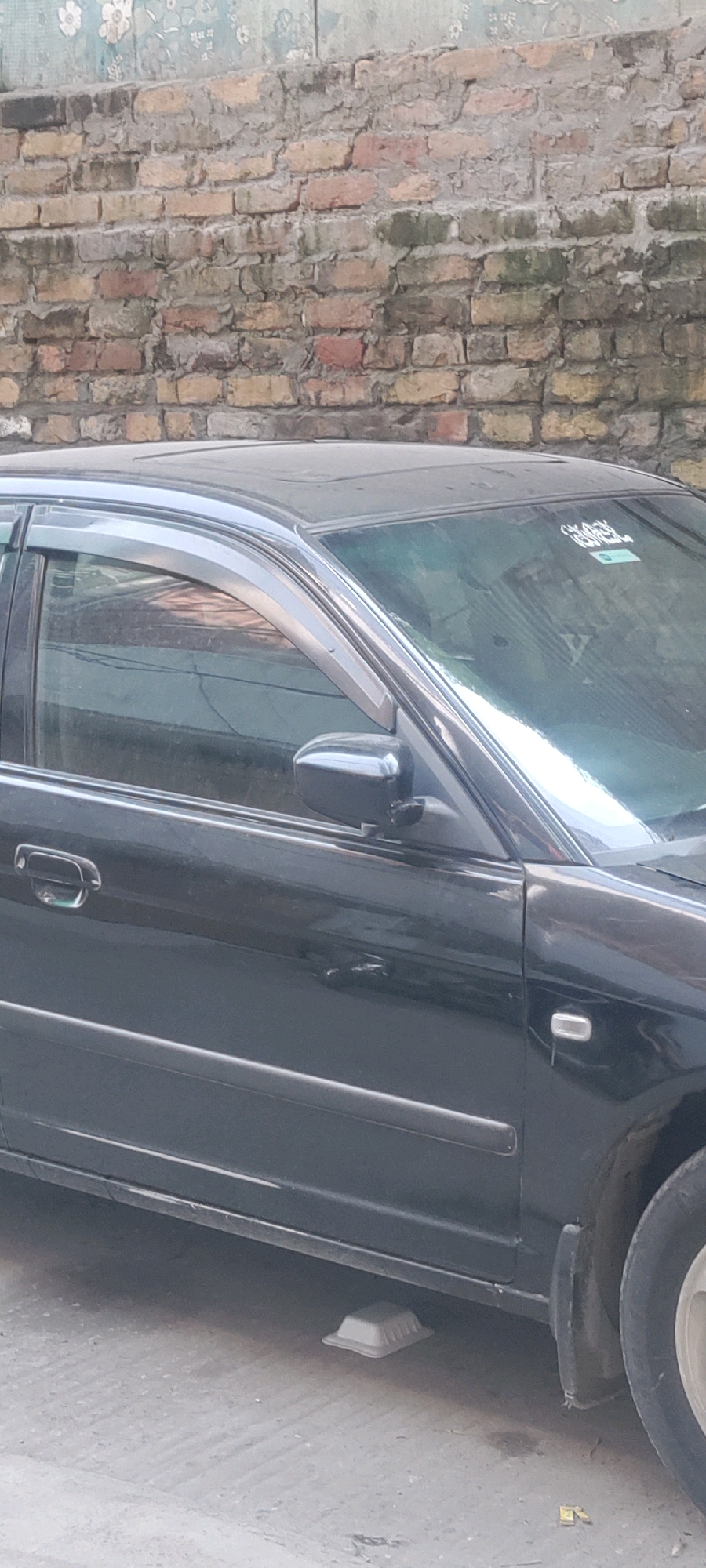 A black sedan is parked on a concrete surface in front of a brick wall. The car has a sunroof and dark tinted windows. A beige plastic container is placed under the front of the car. The brick wall in the background is weathered and shows signs of age. The location is the Metropolitan Corporation Rawalpindi, Pakistan.