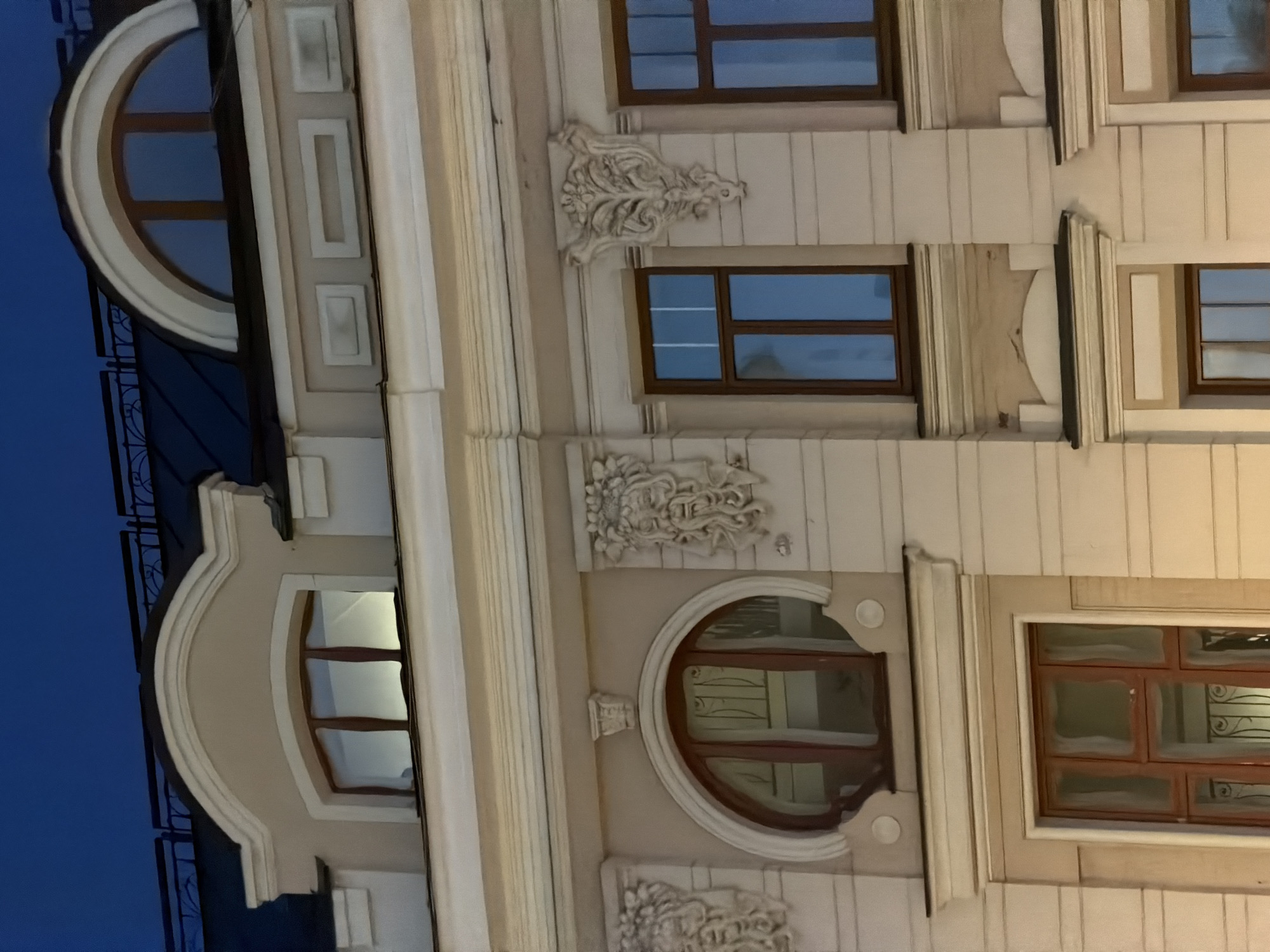 The image depicts a building facade in Moscow, Russia, likely taken at dusk or early evening. The building features an elaborate architectural style with rounded and rectangular windows, ornate moldings, and decorative reliefs. The sky is a deep blue, contrasting with the cream-colored exterior of the building. The windows reflect ambient light, suggesting that the building may be illuminated from within. A wrought-iron balcony can be seen on the far left.