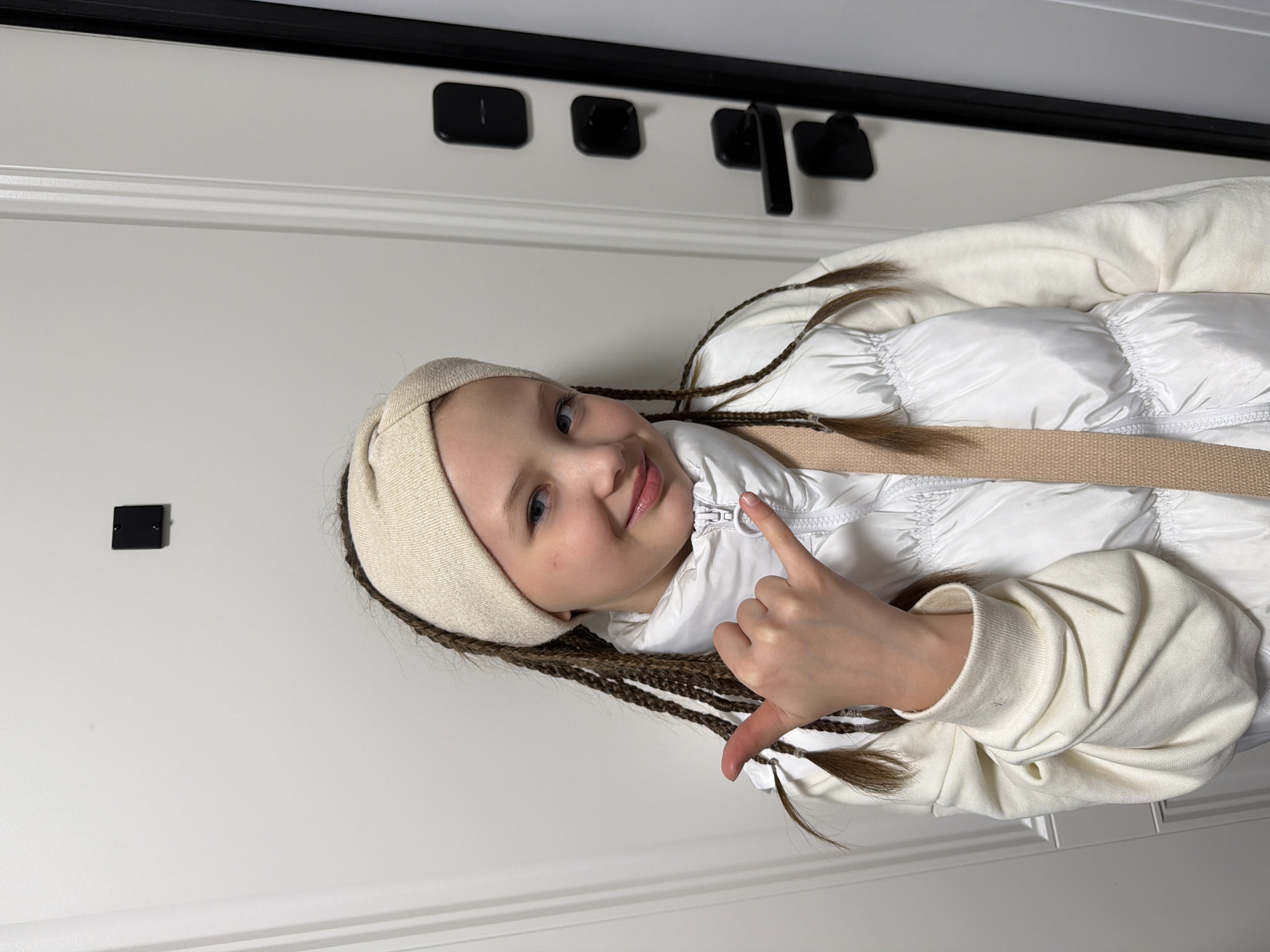 A young girl with long braided hair poses in a white puffer jacket with a tan strap and a matching headband in front of a white door. The door has a black handle, strike plate, and other black rectangular hardware. The location cannot be determined as Chaikovsky, Russia, using the given information.