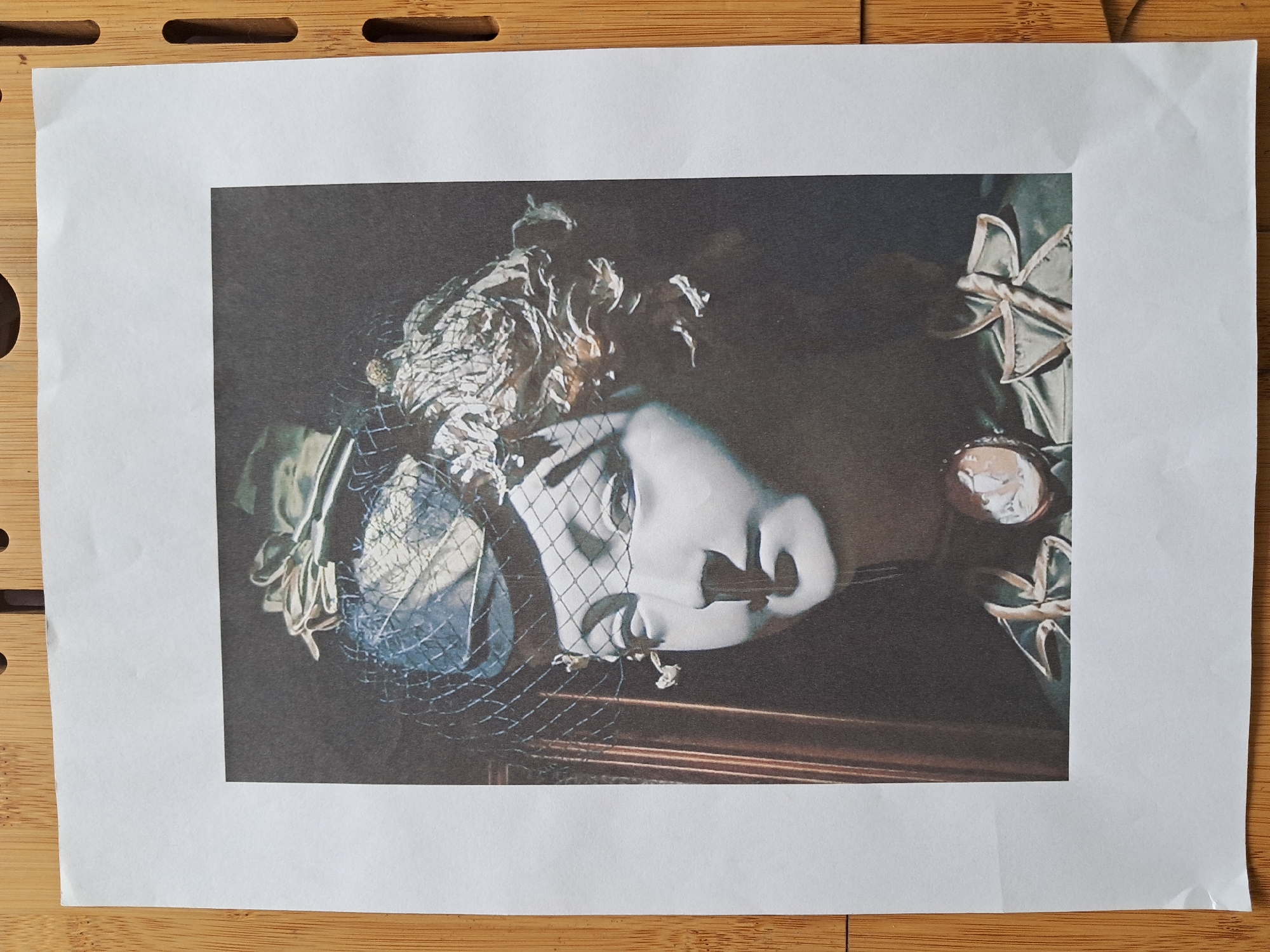 A framed portrait of a mannequin head wearing a hat and veil is displayed against a dark background. The mannequin has delicate features, including closed eyes and a small nose. It is adorned with a cameo brooch at the neck and a decorative ribbon. The hat is embellished with feathers and netting. The portrait is printed on white paper and is slightly creased. The paper is placed on a light brown wooden surface with visible grain and circular indentations.