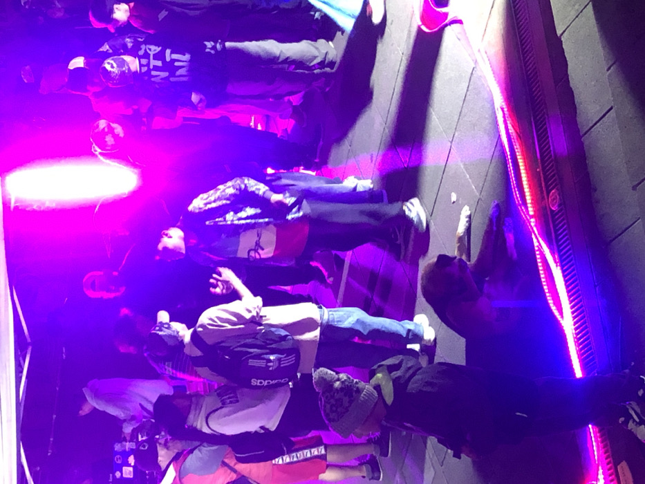 A group of people stand together, illuminated by vibrant pink and purple lights. Most are facing away from the camera, their attire ranging from hoodies and jackets to t-shirts and jeans. Several individuals have backpacks or hats. The scene seems to be outdoors, with a wall featuring embedded lights visible in the background. The lighting creates long shadows and a dynamic atmosphere.
