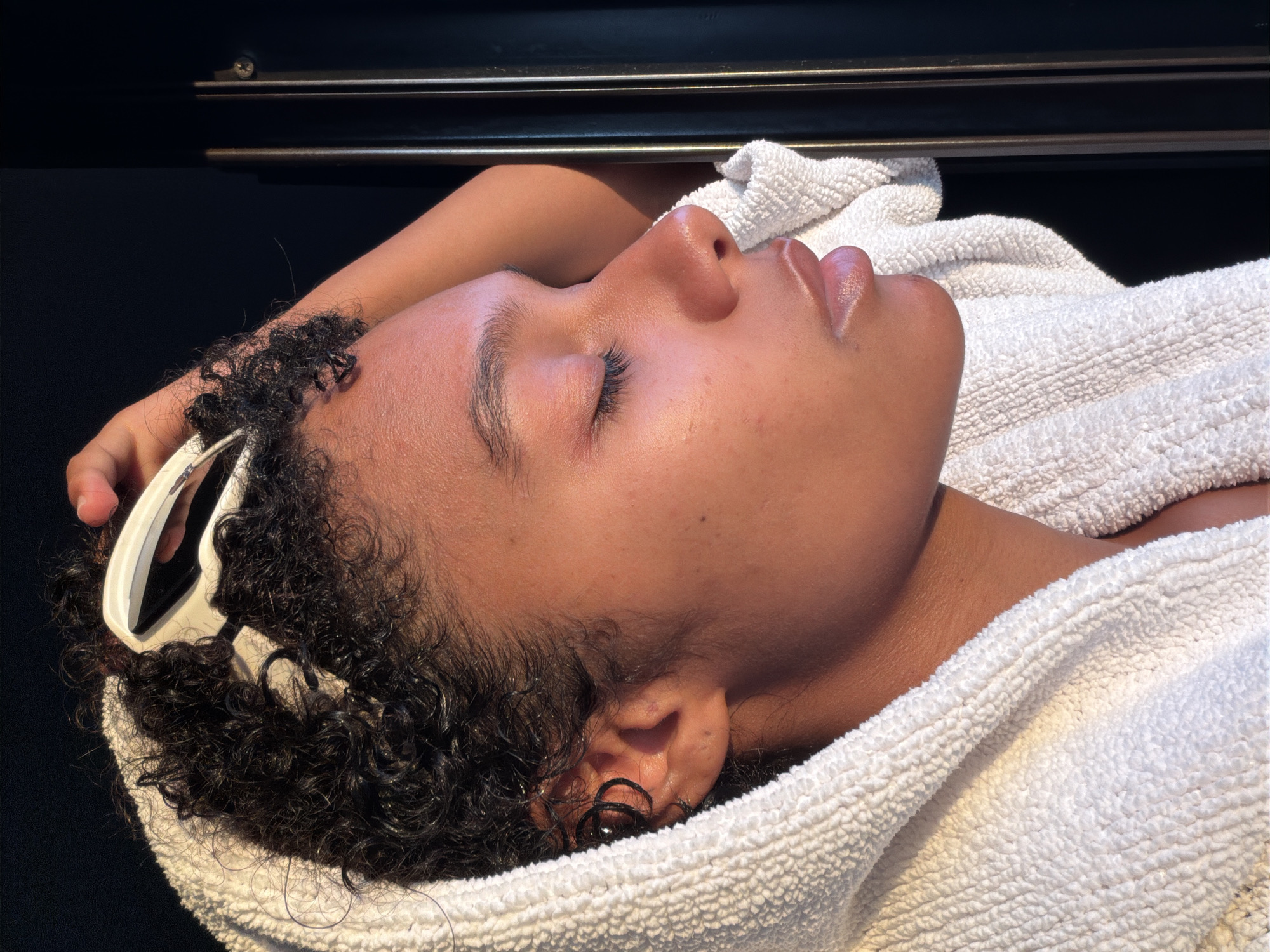 The image is a close-up, side profile of a young person with dark, curly hair and closed eyes, wrapped in a white towel. They are holding a white hair styling tool to their hair. The lighting suggests a natural, possibly indoor setting with warm tones illuminating the subject's skin. The background is dark and slightly out of focus, with vertical lines visible on the right side, possibly indicating a door or a reflective surface. The person is positioned in a relaxed, contemplative manner, with their head tilted back and arm resting behind them. There is no identifiable connection to Champs-sur-Marne, France, within the visual information provided in the image.